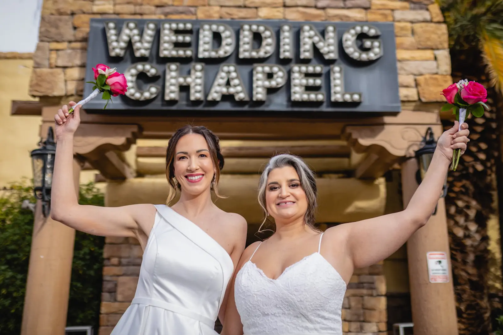 Two brides holding pink rose bouquets celebrating outside the Las Vegas Wedding Chapel marquee sign