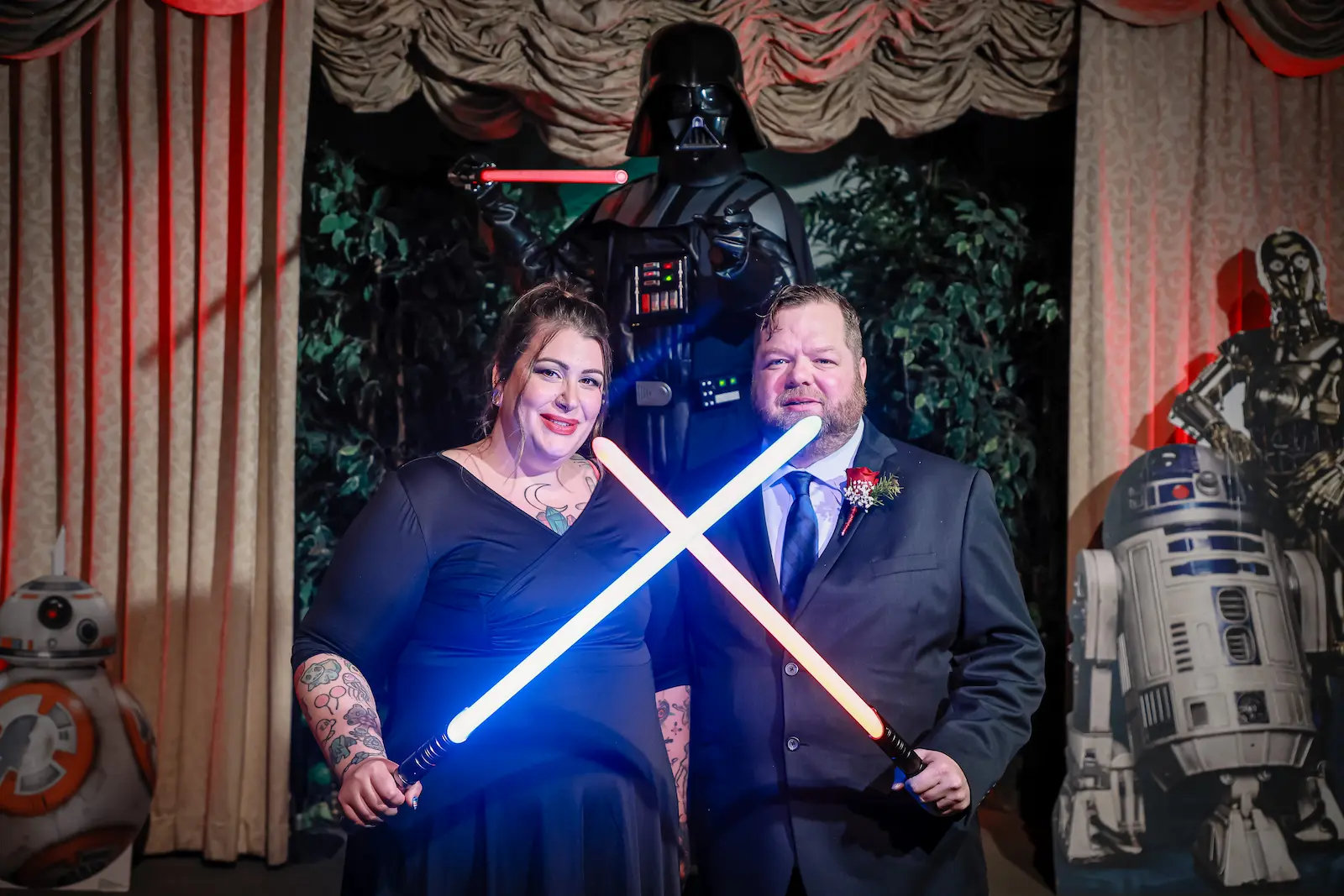 Couple crossing blue and red lightsabers with Darth Vader and droid props on Star Wars themed stage