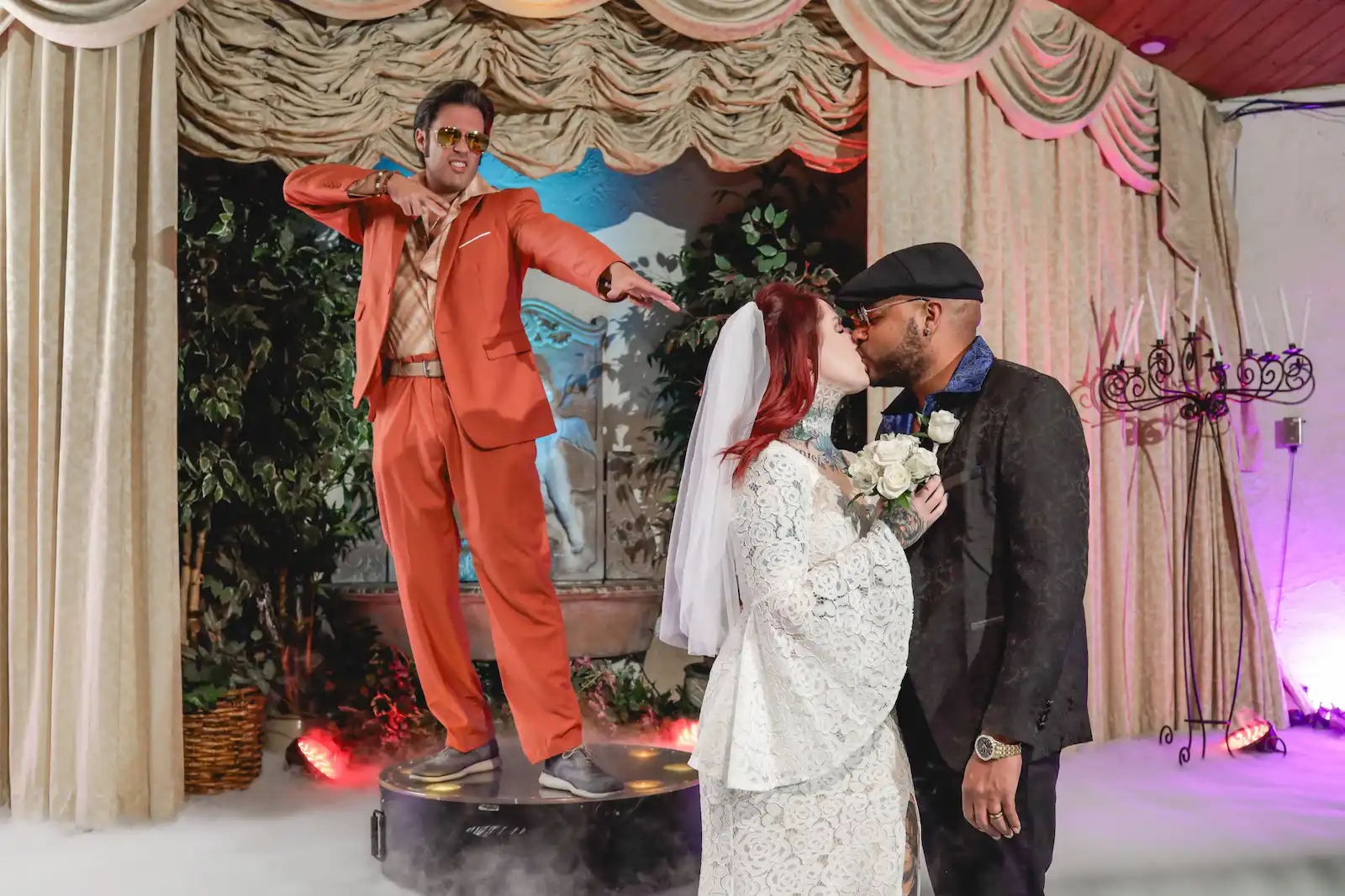 Couple sharing their first kiss while Elvis impersonator in orange suit points from stage with fog at Las Vegas chapel