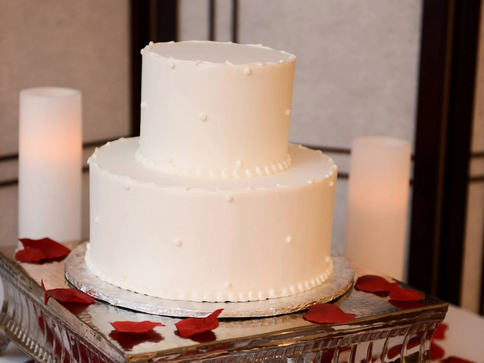 Two-tier white wedding cake with pearl dot details surrounded by red rose petals and candles