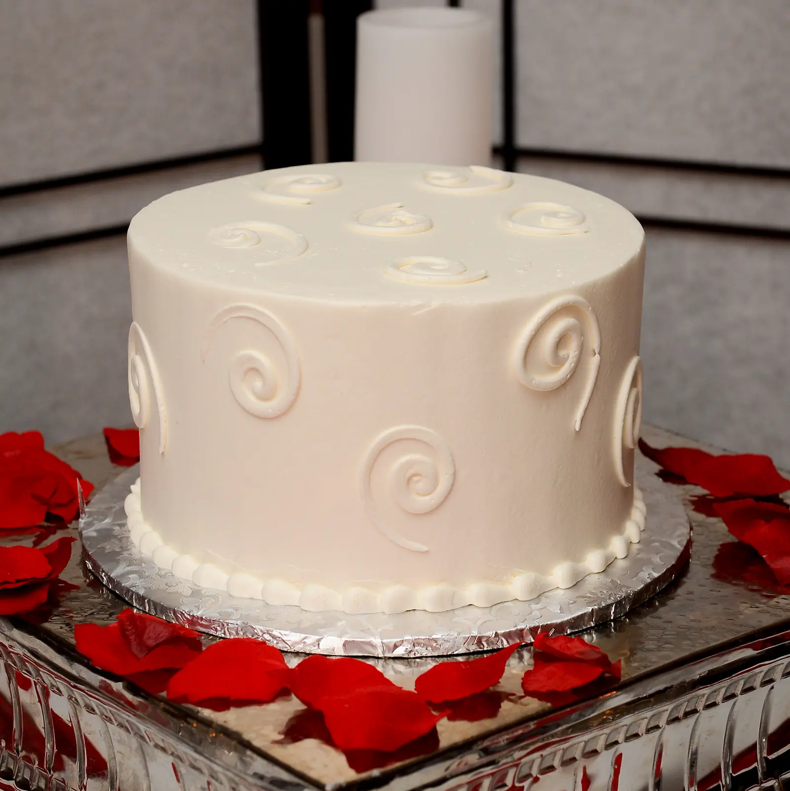 Single tier white wedding cake with swirl design on a silver tray with red rose petals