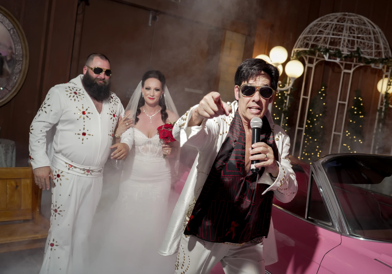 Groom in an Elvis jumpsuit and bride pose as an Elvis impersonator sings next to a pink Cadillac