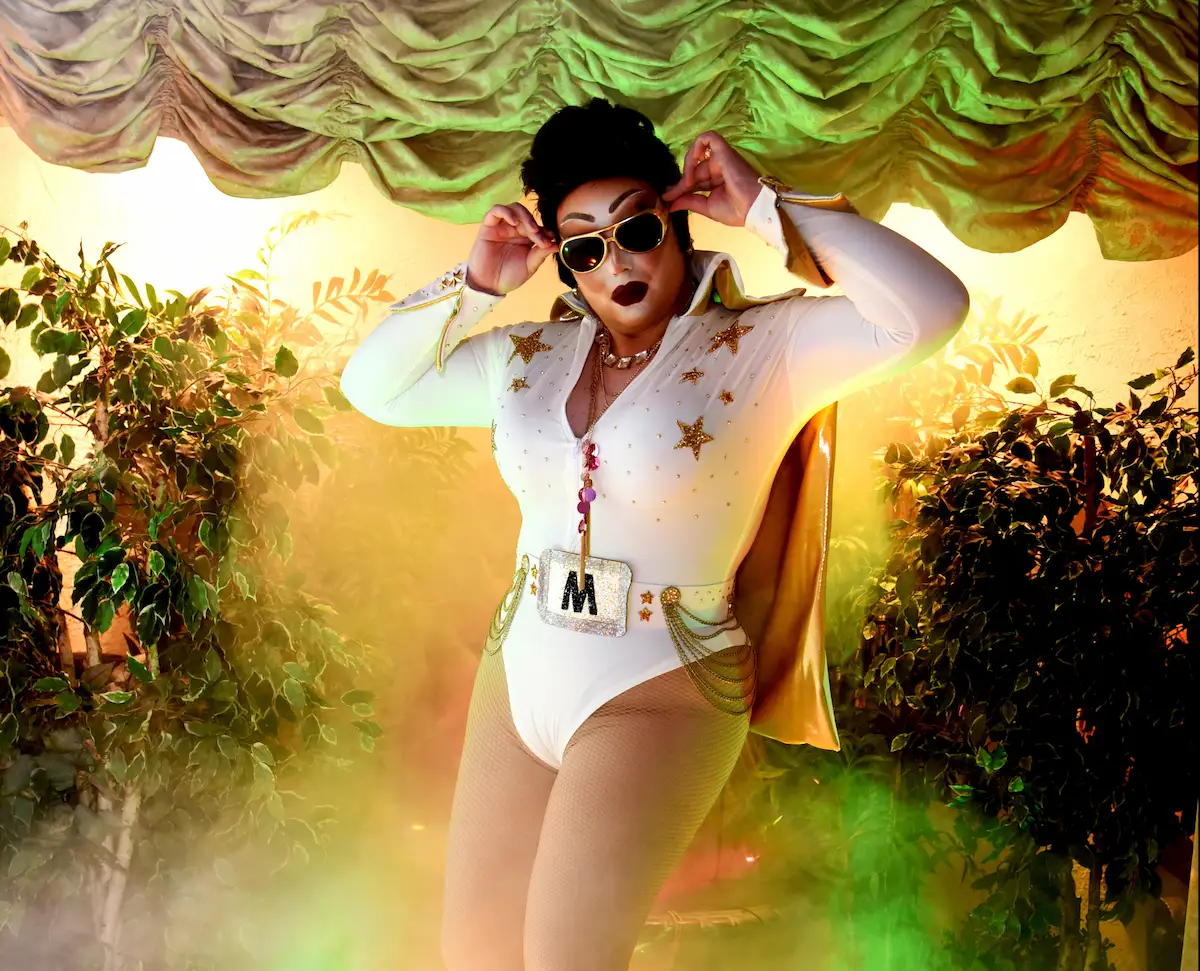 Drag Elvis in a white jumpsuit and gold cape poses on stage at a Vegas chapel