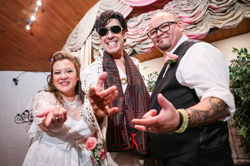 Bride and groom posing with an Elvis impersonator in a white jumpsuit at a Las Vegas Elvis wedding chapel.