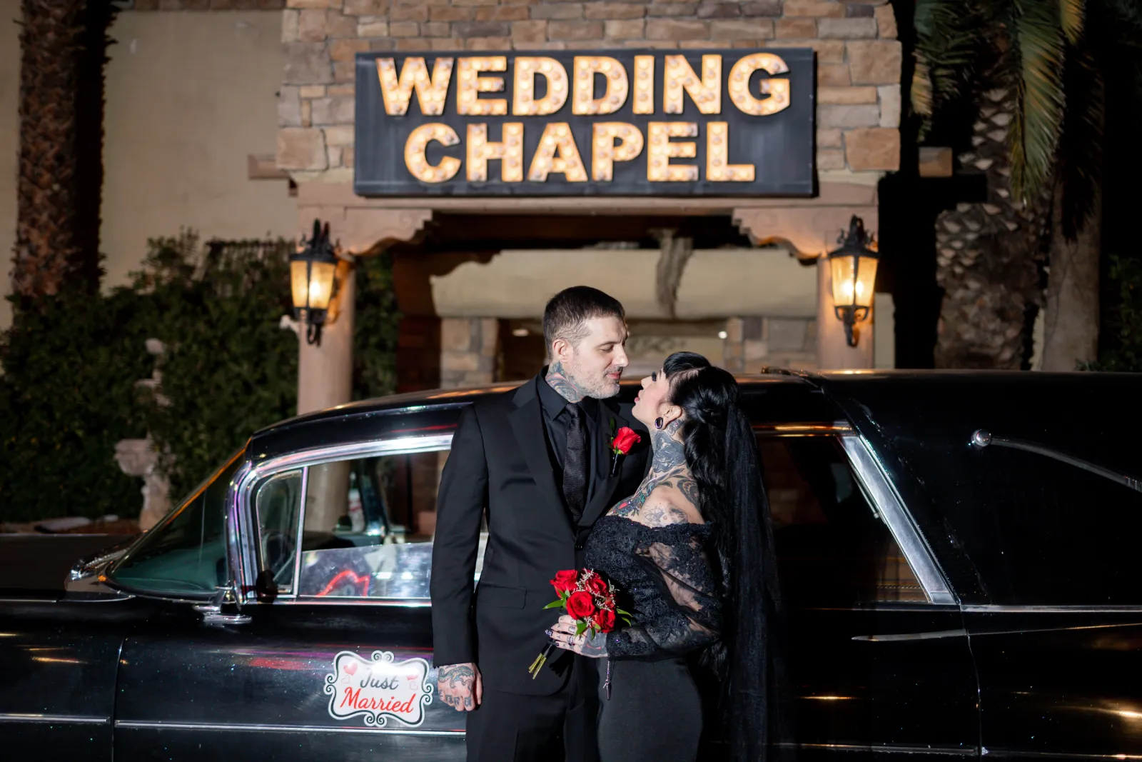 Tattooed gothic couple beside a vintage black car under a lit Wedding Chapel marquee in Las Vegas