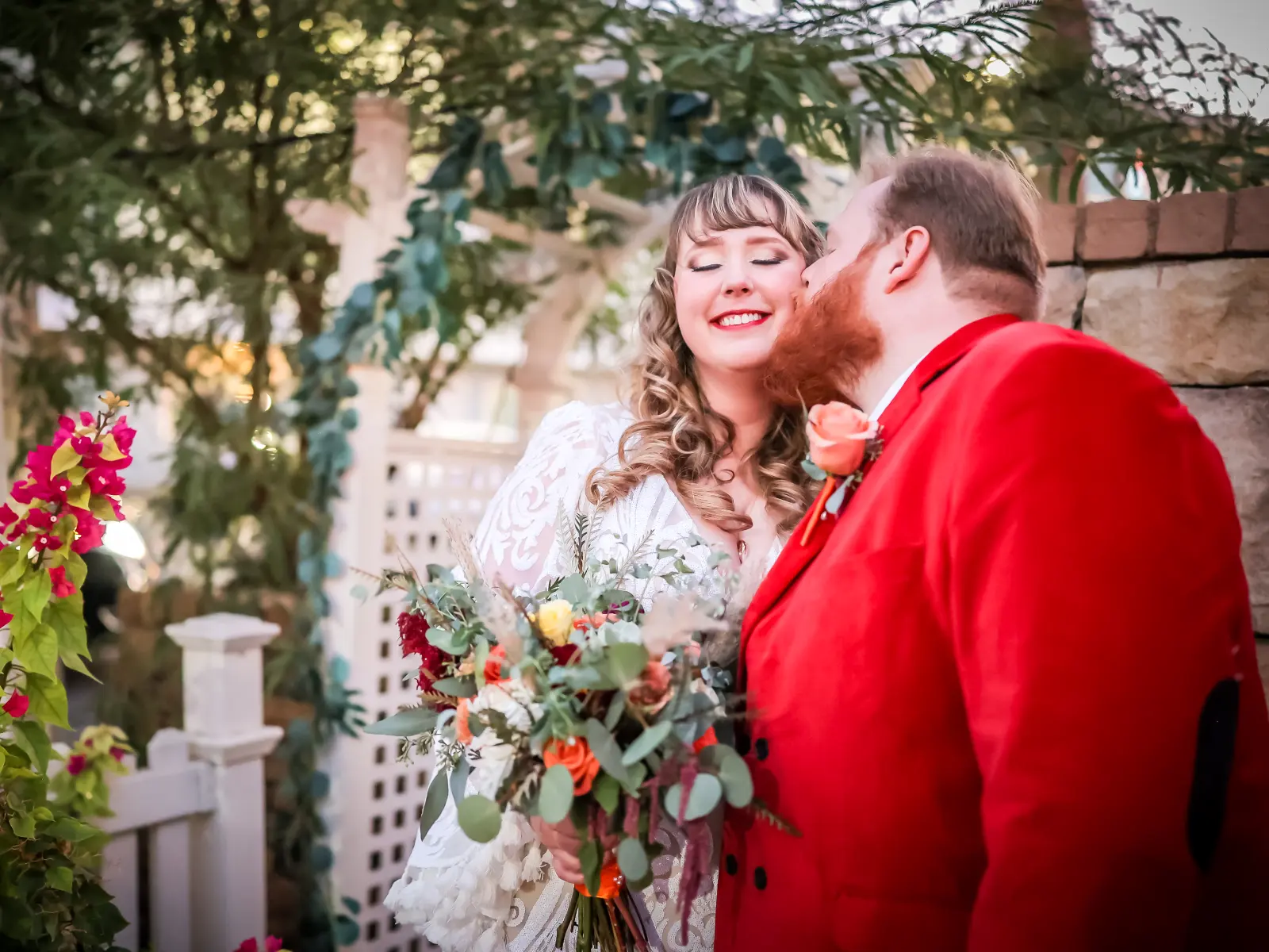 A groom in a bright red suit kisses a smiling bride
