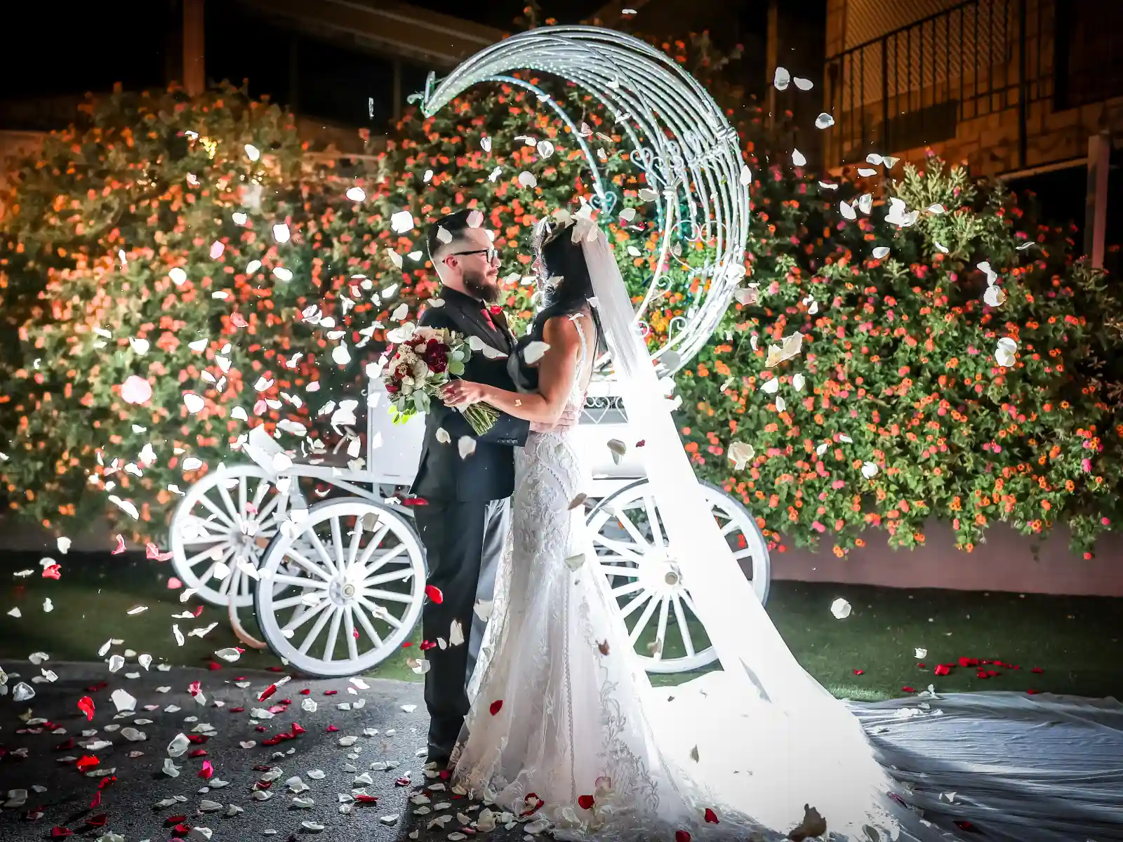 A bride and groom embrace outdoors at night while white and red petals fall around them, with a white carriage and flower bushes in the background
