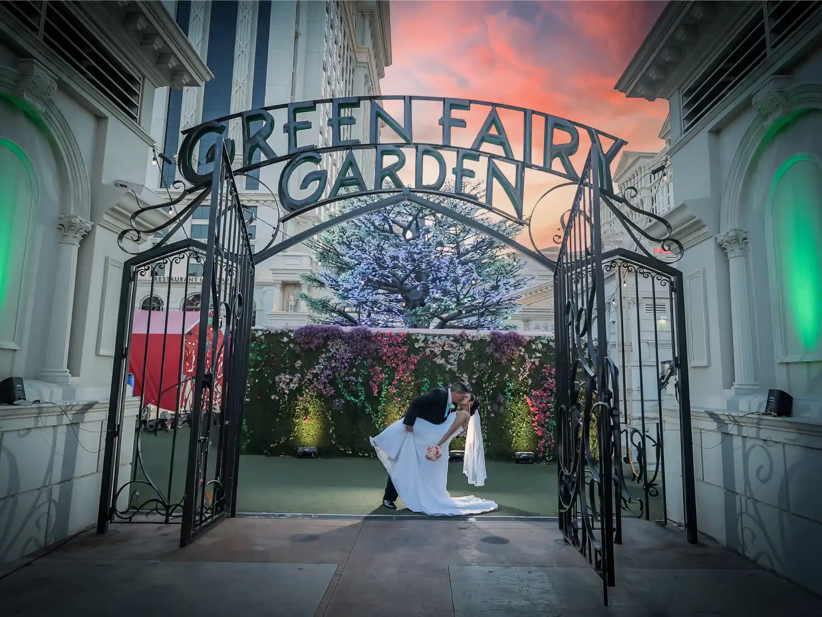 A bride and groom kiss on a small bridge leading to an ornate white gazebo filled with flowers and greenery inside a large conservatory