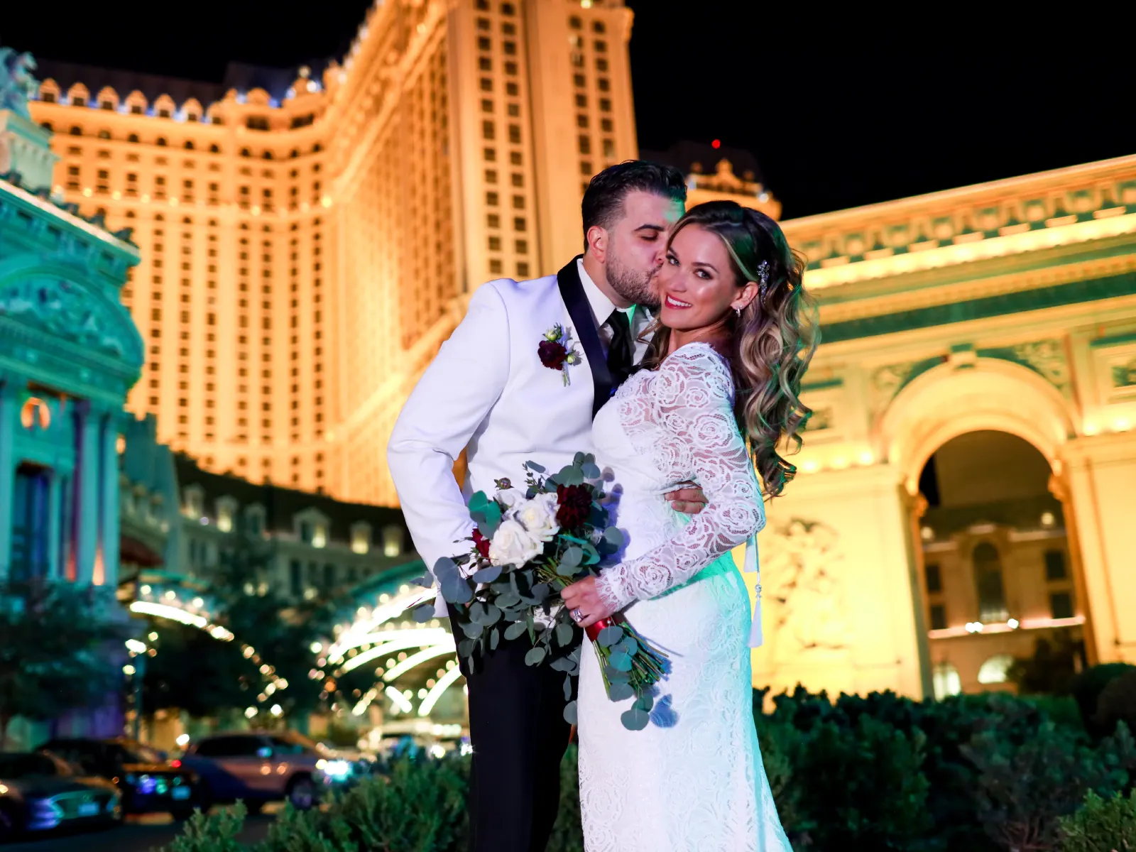 A groom in a white tuxedo kisses a bride
