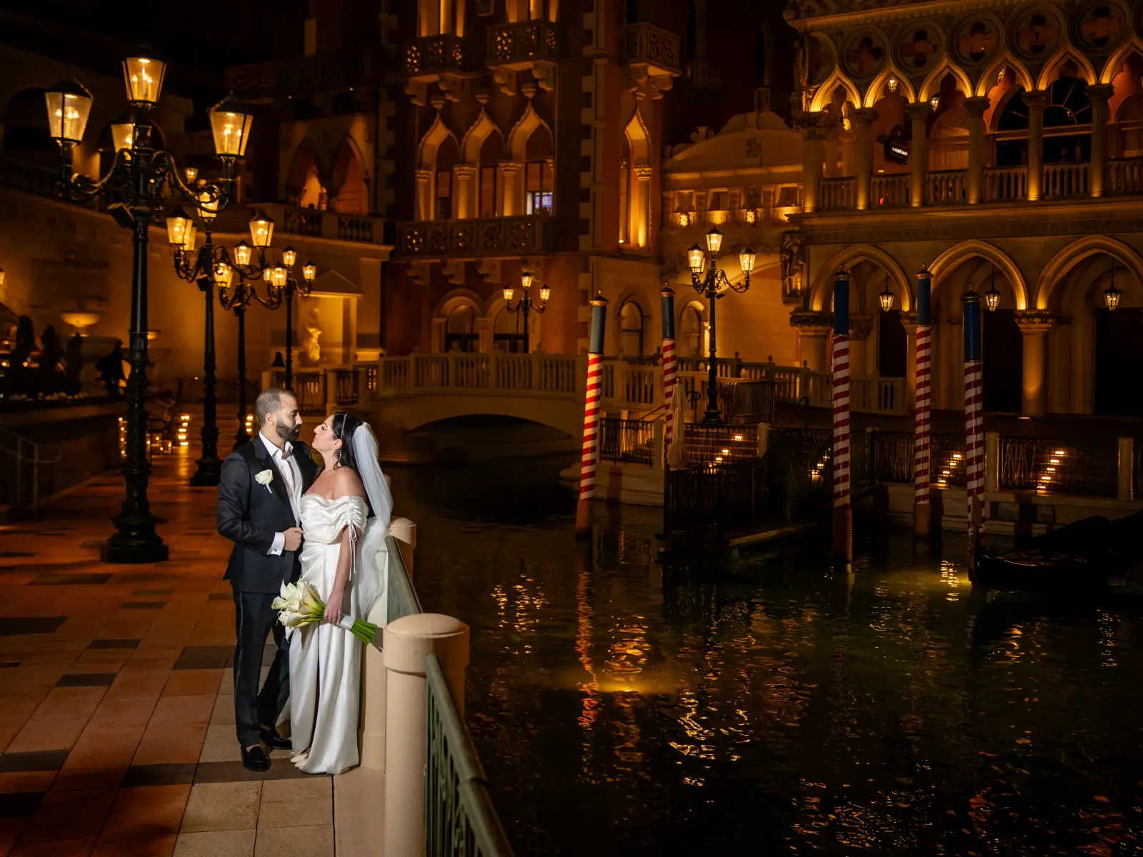 A bride and groom stand by the Venetian canal in Las Vegas at night, with Italian-style architecture, striped gondola poles, and glowing street lamps in the background.
