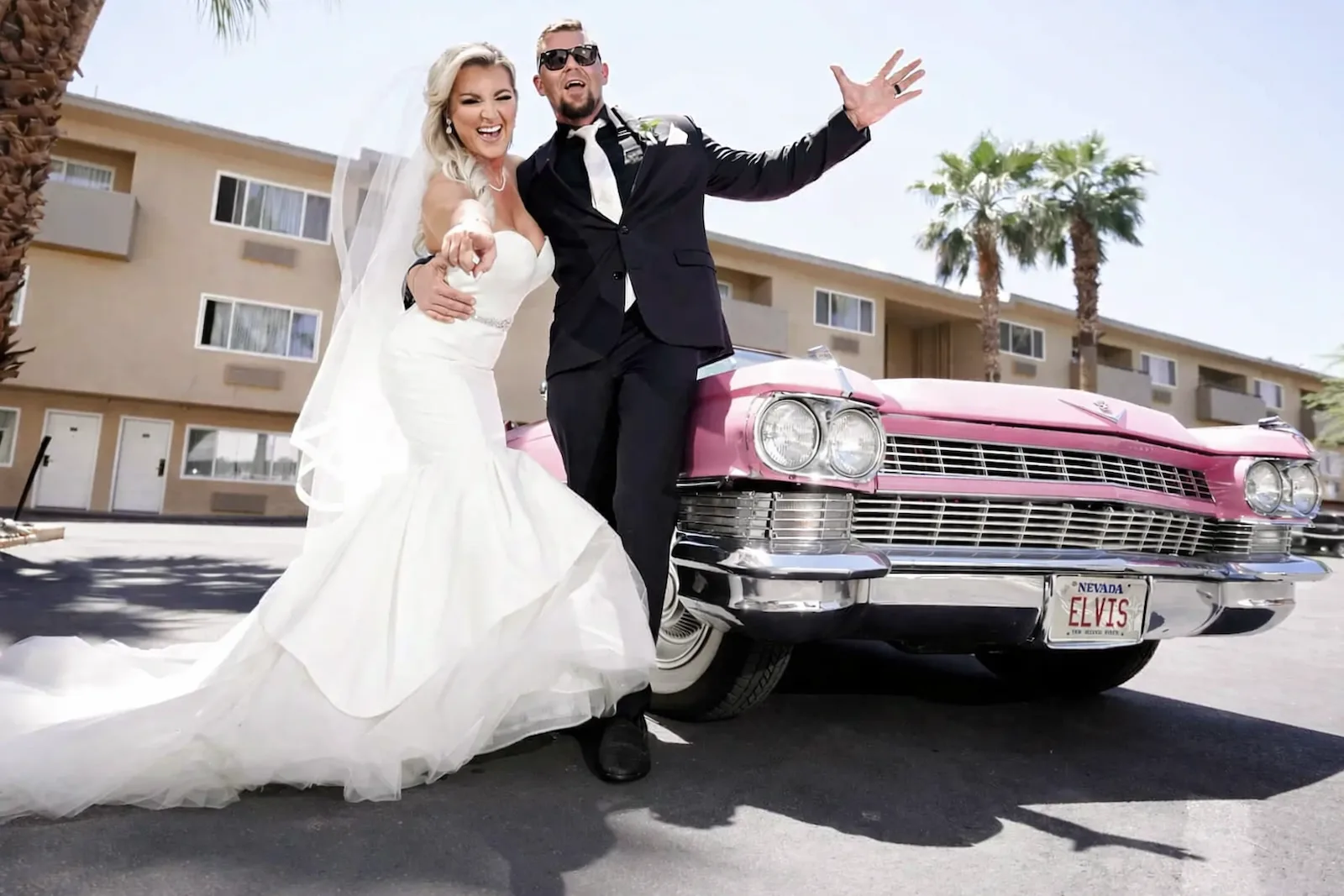 Newlywed couple posing with a vintage pink Cadillac with Nevada Elvis license plate outside a Las Vegas wedding chapel with palm trees