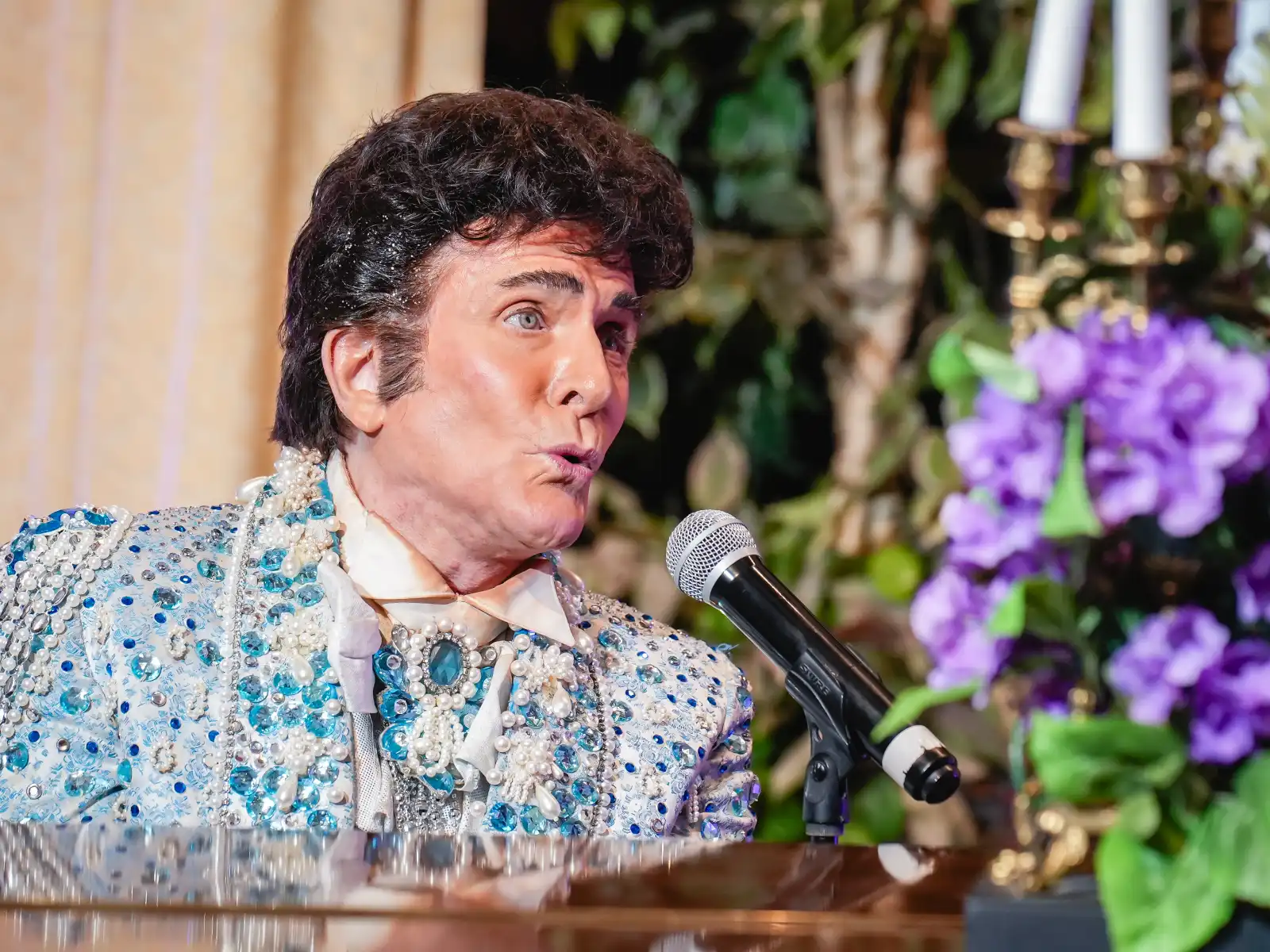 A close-up profile of a Liberace impersonator with voluminous dark hair, wearing a high-collared blue suit heavily embellished with silver beads and pearls, singing into a microphone at a piano.