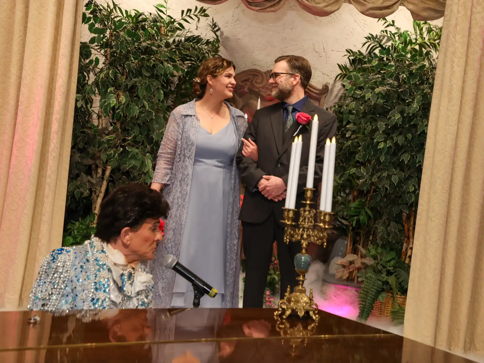 A bride and groom stand at the altar during a ceremony, framed by a candelabra in the foreground and a Liberace tribute performer in a sparkling blue suit seated at the piano in the lower left.