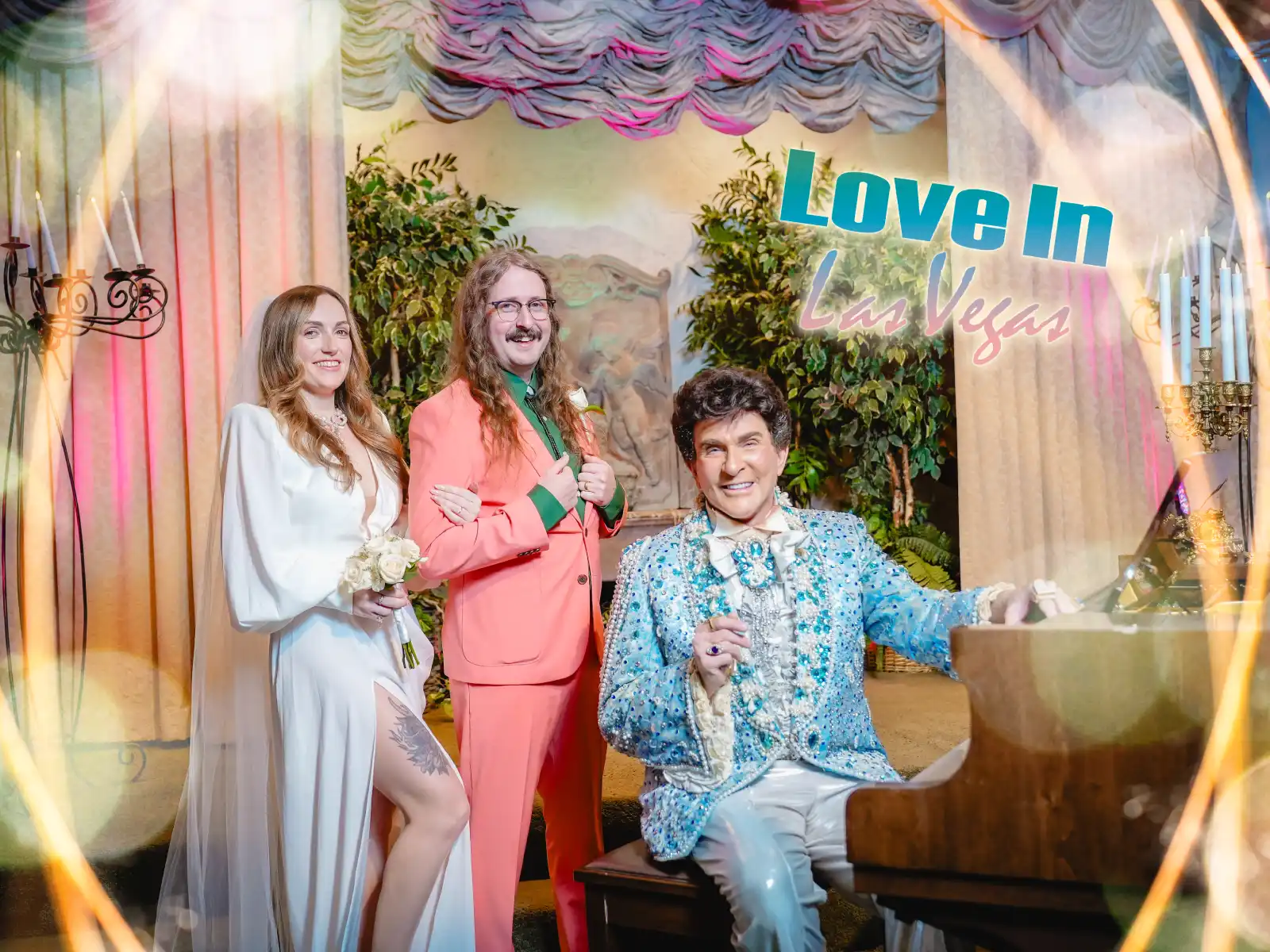 A bride in a white gown and a groom in an orange suit stand behind a Liberace performer seated at a grand piano. A graphic overlay reads "Love In Las Vegas" in a retro font.