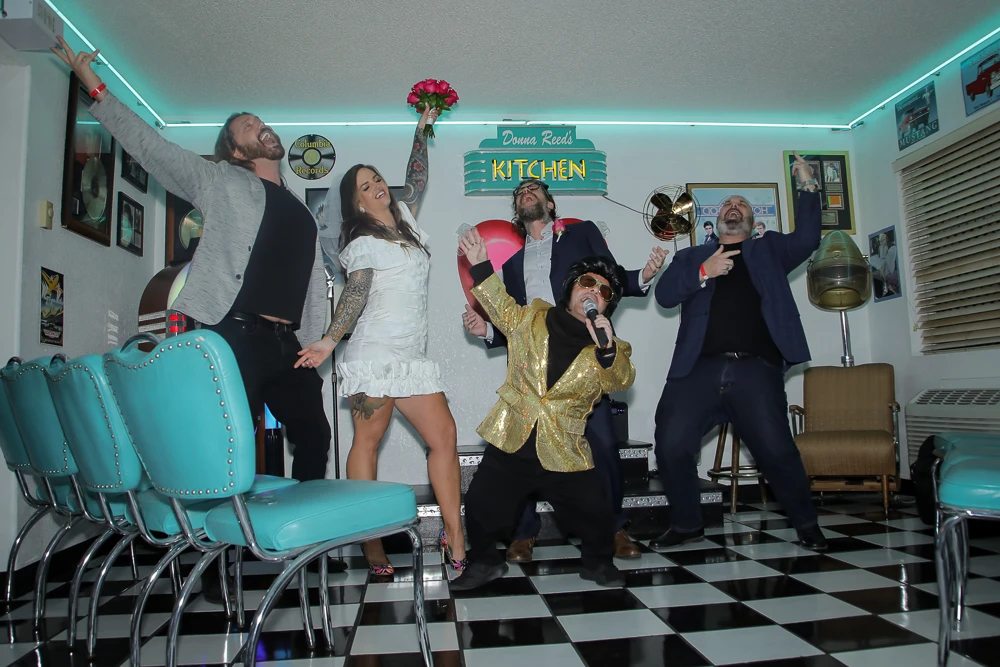 Fun Las Vegas Elvis wedding group photo with bride, groom, friends and Elvis impersonator singing together inside retro diner chapel with checkered floor and neon decor