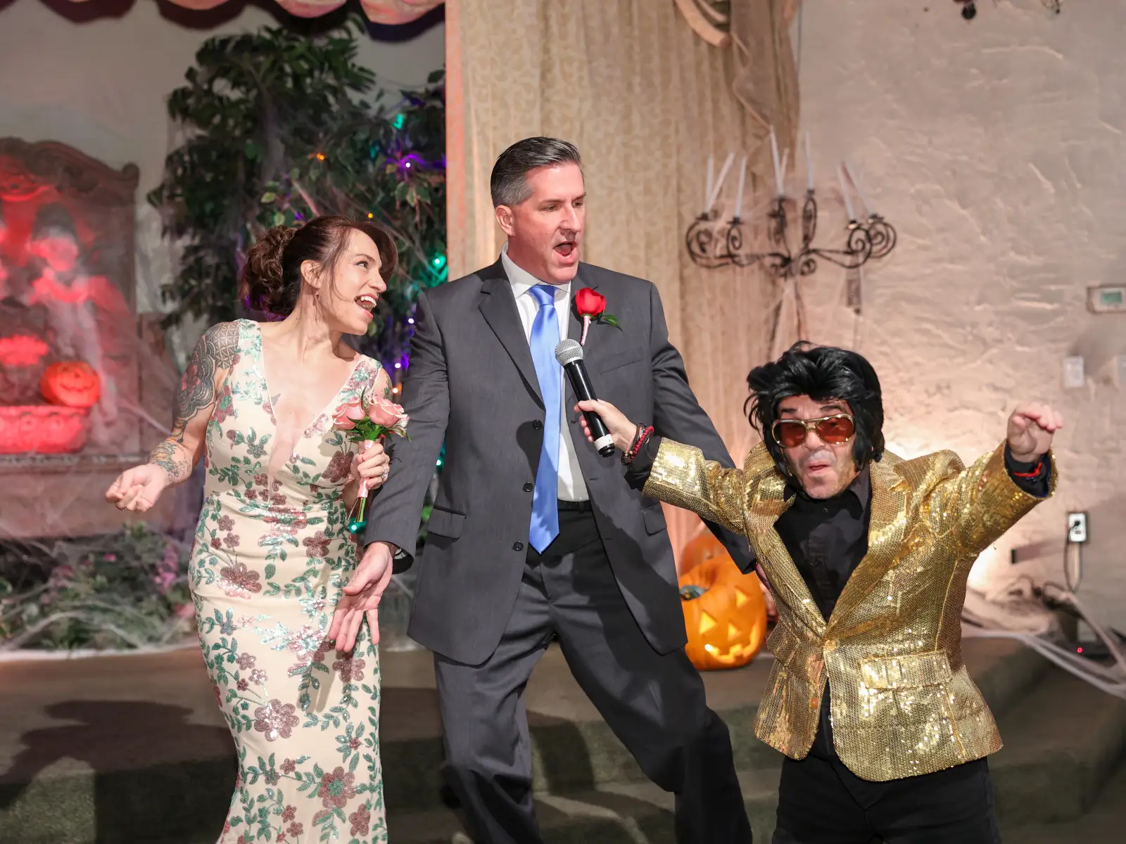 Playful Las Vegas Elvis wedding ceremony moment as lively Elvis impersonator holds microphone toward excited groom while smiling bride in floral dress laughs during festive chapel celebration
