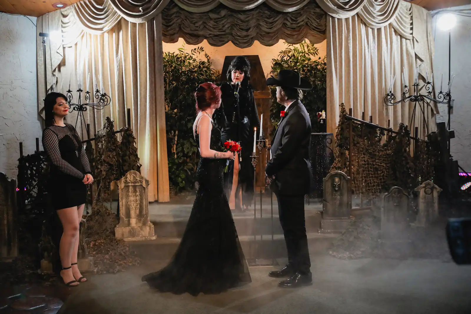Gothic horror hostess impersonator officiating wedding ceremony as bride in black gown and groom exchange vows on graveyard-decorated stage at Las Vegas wedding chapel