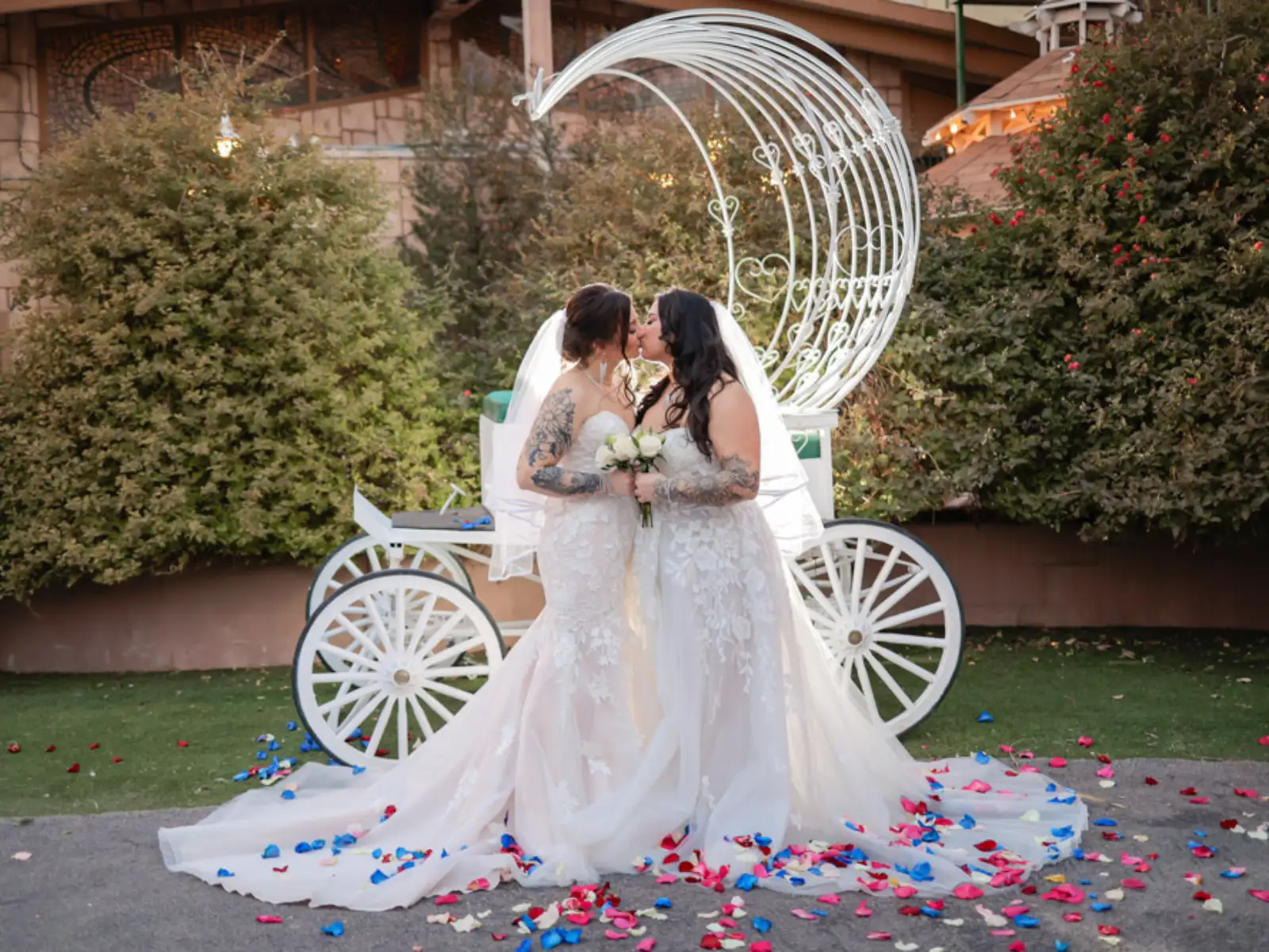 Two brides kissing in front of white fairytale carriage with rose petals at a Las Vegas wedding chapel