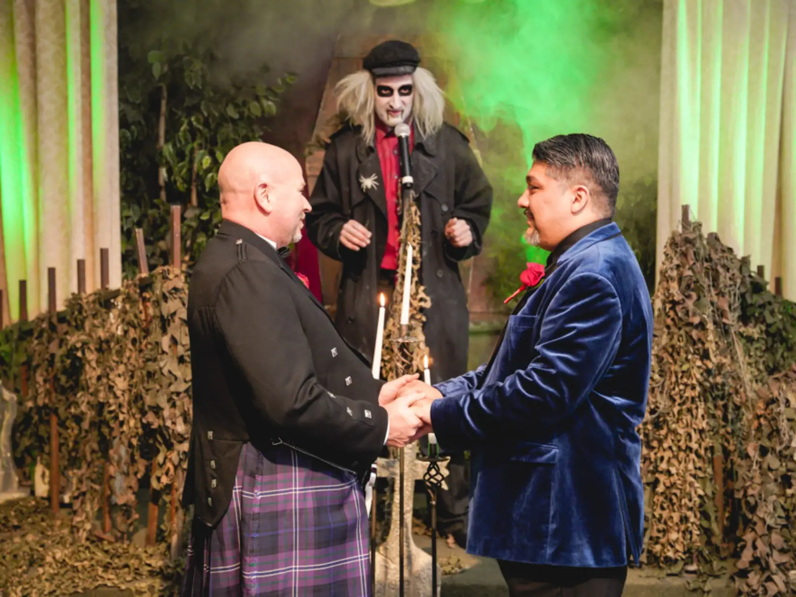 Two grooms holding hands during a Las Vegas gothic wedding ceremony with a Beetlejuice character officiating behind green fog.