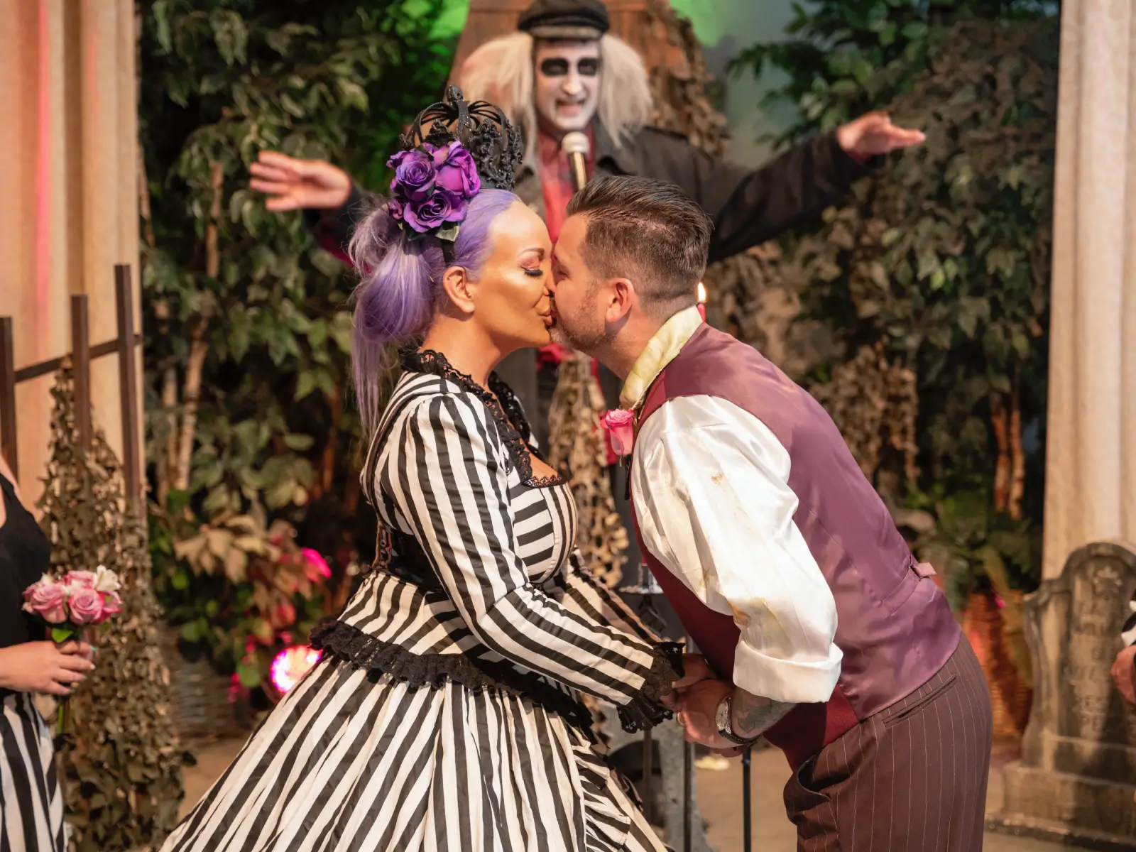 Couple kissing during Beetlejuice-themed wedding ceremony with officiant behind them on stage.