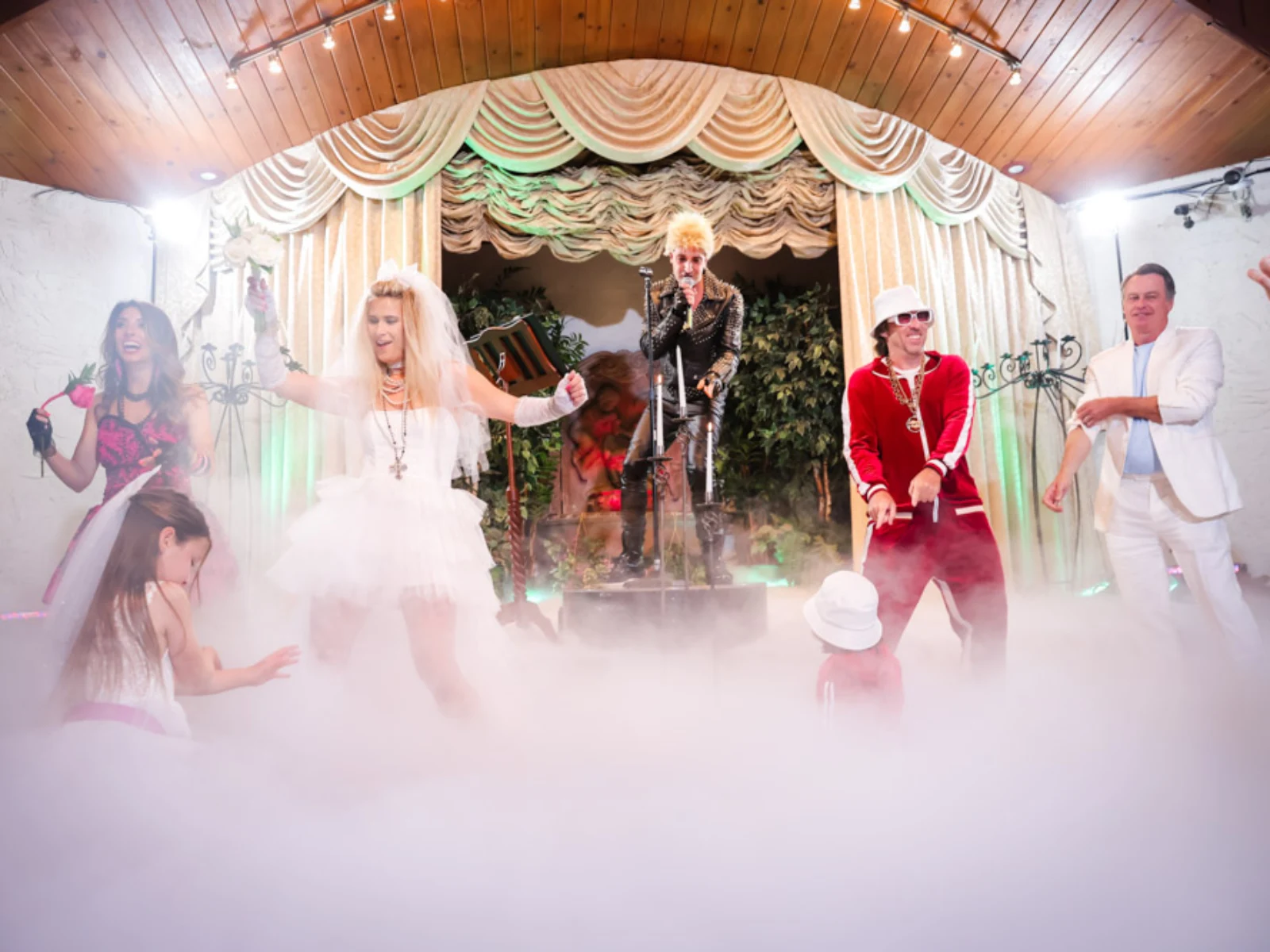 A lively wedding reception scene featuring a Billy Idol impersonator performing on a fog-filled stage. A bride in a short tutu-style dress and white veil dances in the foreground alongside guests, including a man in a red tracksuit and a flower girl, under warm stage lighting.