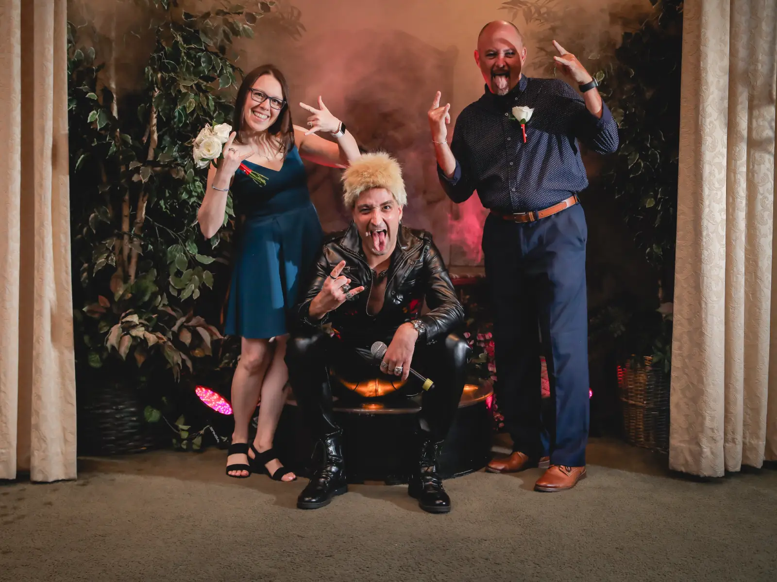 A Billy Idol impersonator in a studded leather jacket and blonde spiked hair sits on a stage, posing with a rock-and-roll hand gesture and tongue out. He is flanked by two wedding guests in formal attire who are also making rock hand signs against a backdrop of indoor greenery and curtains.