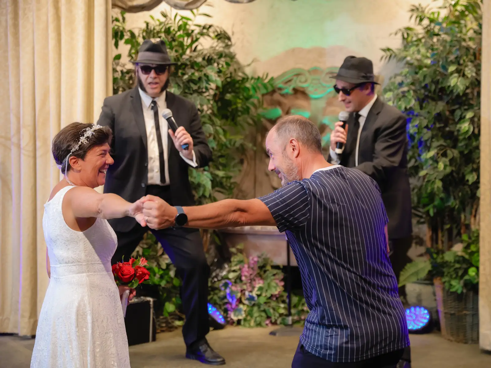 A laughing bride in a white lace dress dances with a guest while two Blues Brothers impersonators perform in the background.