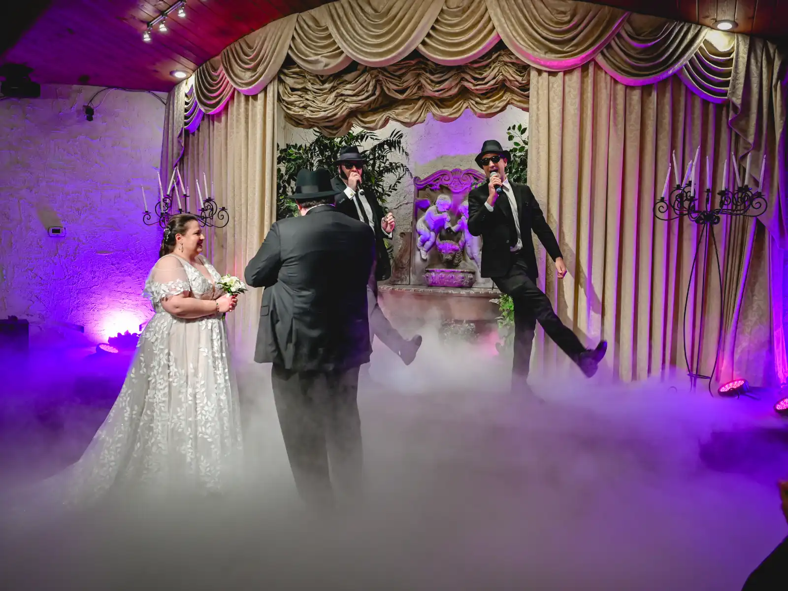 A wedding couple stands on a fog-filled dance floor as two Blues Brothers tribute performers dance and sing under stage lights.