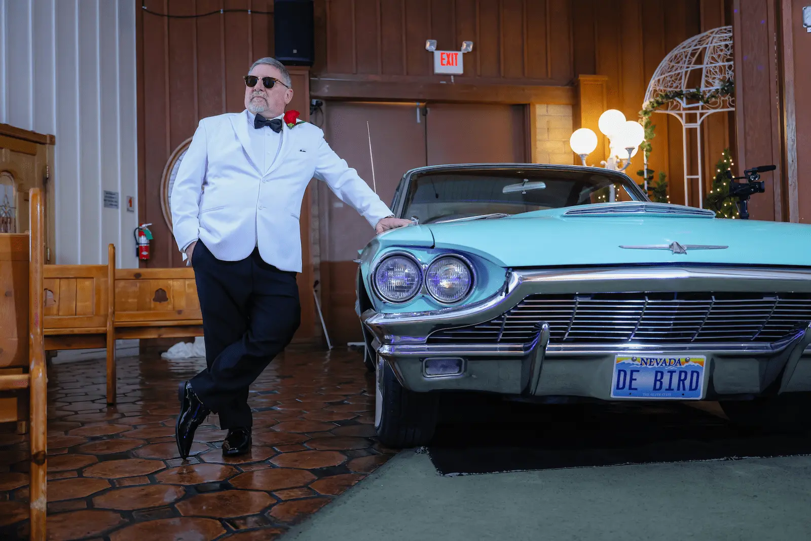 Groom in white tuxedo leans against vintage turquoise Thunderbird inside James Bond themed Las Vegas wedding chapel