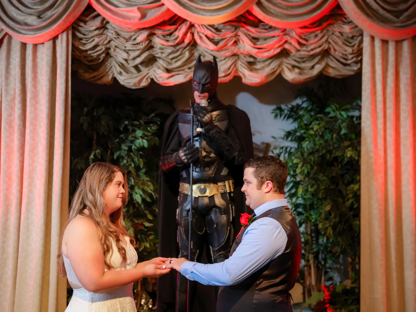 A bride in a white lace dress and a groom in a blue shirt and black vest exchange vows. Behind them, a person in a realistic Batman costume speaks into a microphone under warm red stage lighting.