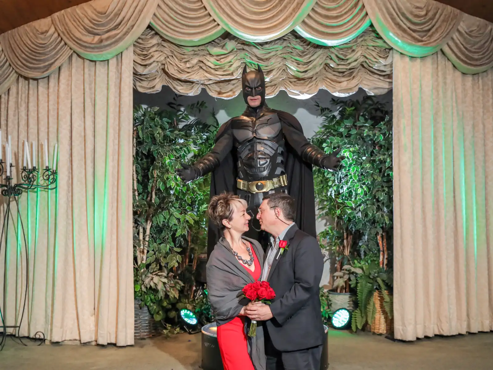 A couple in formal attire, the woman in a red dress and grey shawl, stands before a life-sized Batman figure acting as a wedding officiant. The stage is decorated with beige curtains and green-tinted lighting.