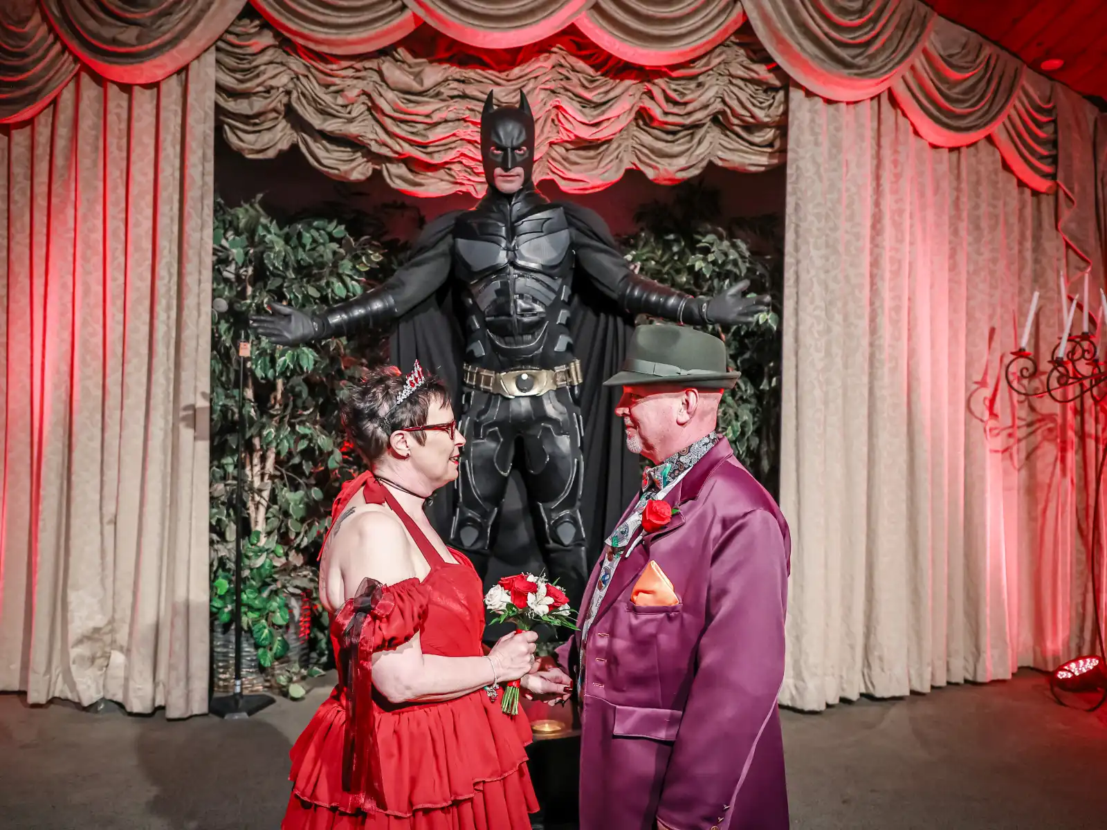 A bride in a ruffled red dress and a groom in a purple suit and fedora face each other at the altar. A life-sized Batman statue stands behind them with arms outstretched under vibrant red theatrical lighting.