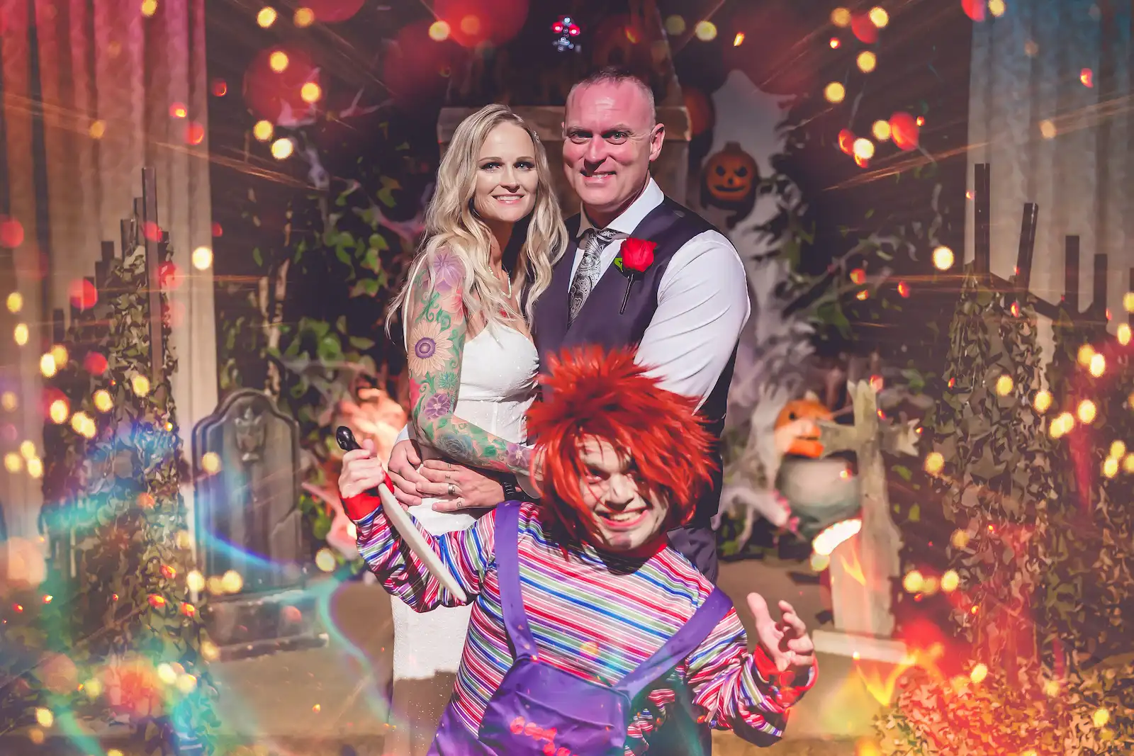 Tattooed couple smiling with a Chucky character surrounded by bokeh lights at a Las Vegas halloween wedding.