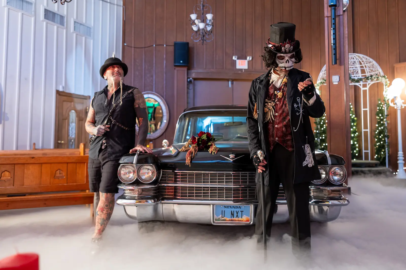 Voodoo skeleton character and tattooed performer posing in front of a vintage black hearse with fog inside a Las Vegas wedding chapel.