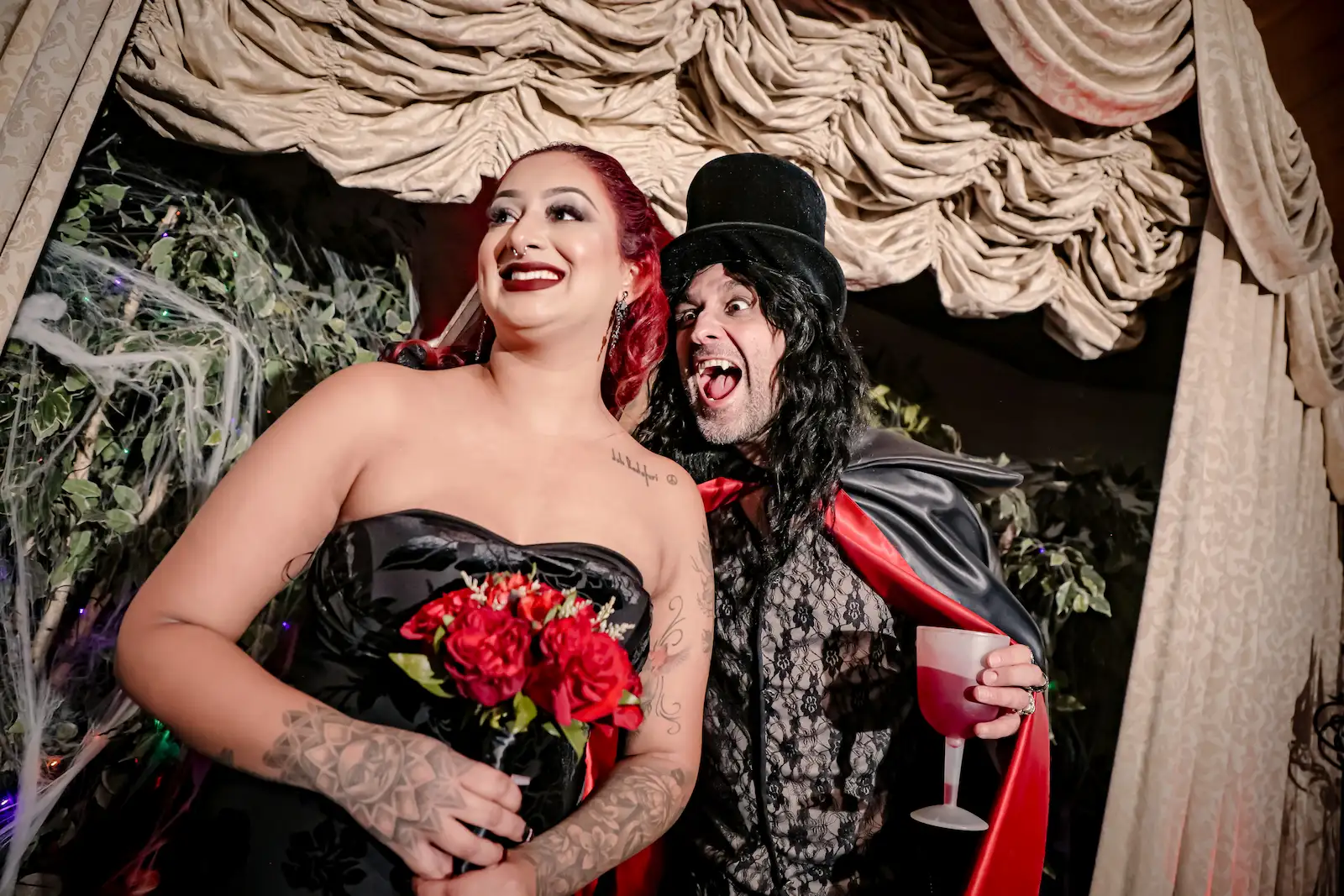 Gothic bride in a black dress with red roses posing with a vampire character at a Las Vegas horror wedding.