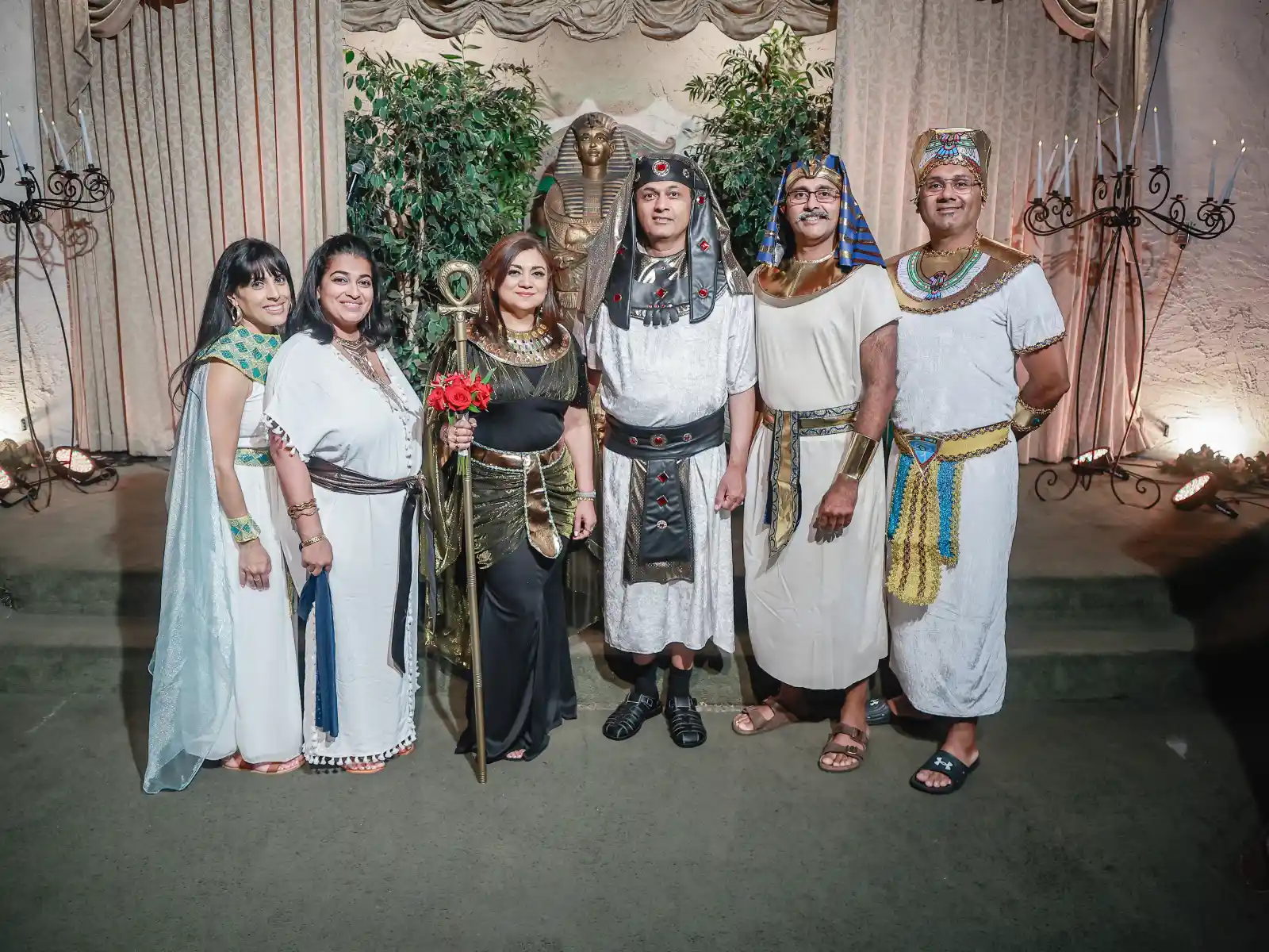 A group of six people posing in various Ancient Egyptian inspired costumes, including tunics, ornate collars, and headdresses, in front of a sarcophagus prop.