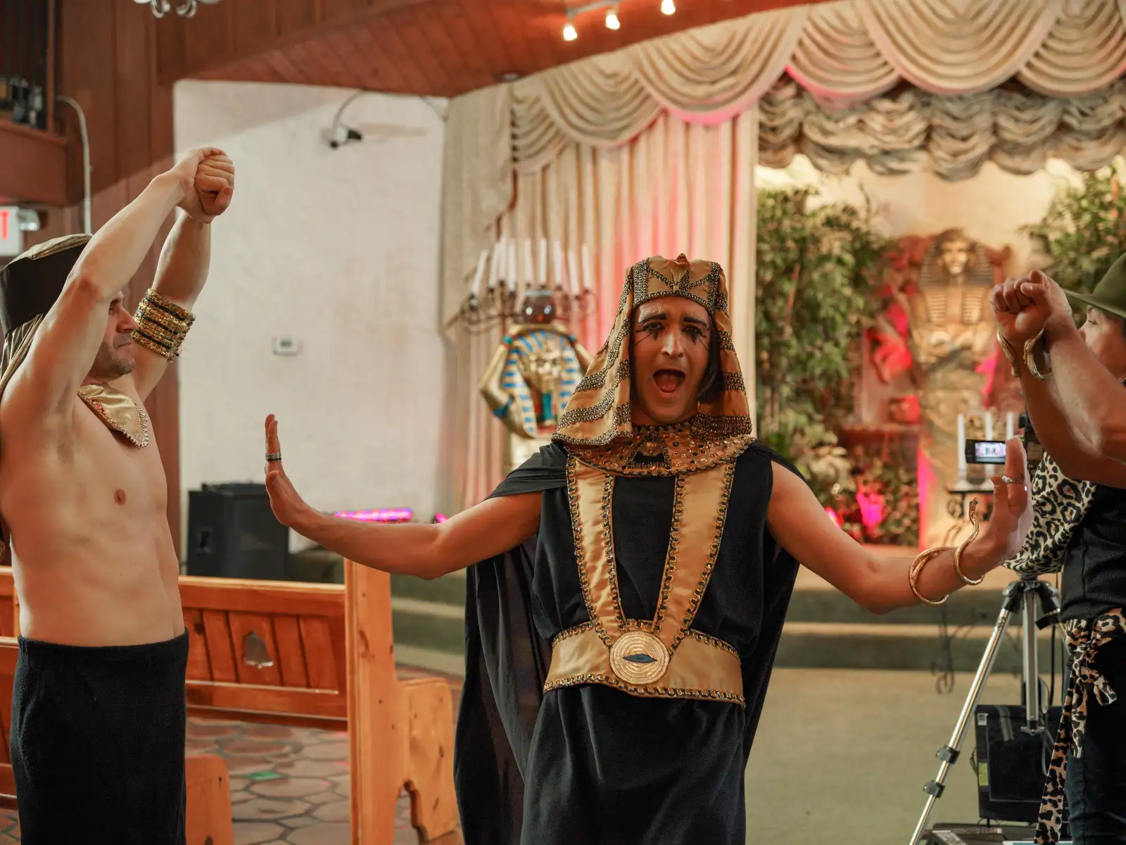 A performer in a black and gold Egyptian tunic and headdress strikes a dramatic pose with arms outstretched during a themed event.