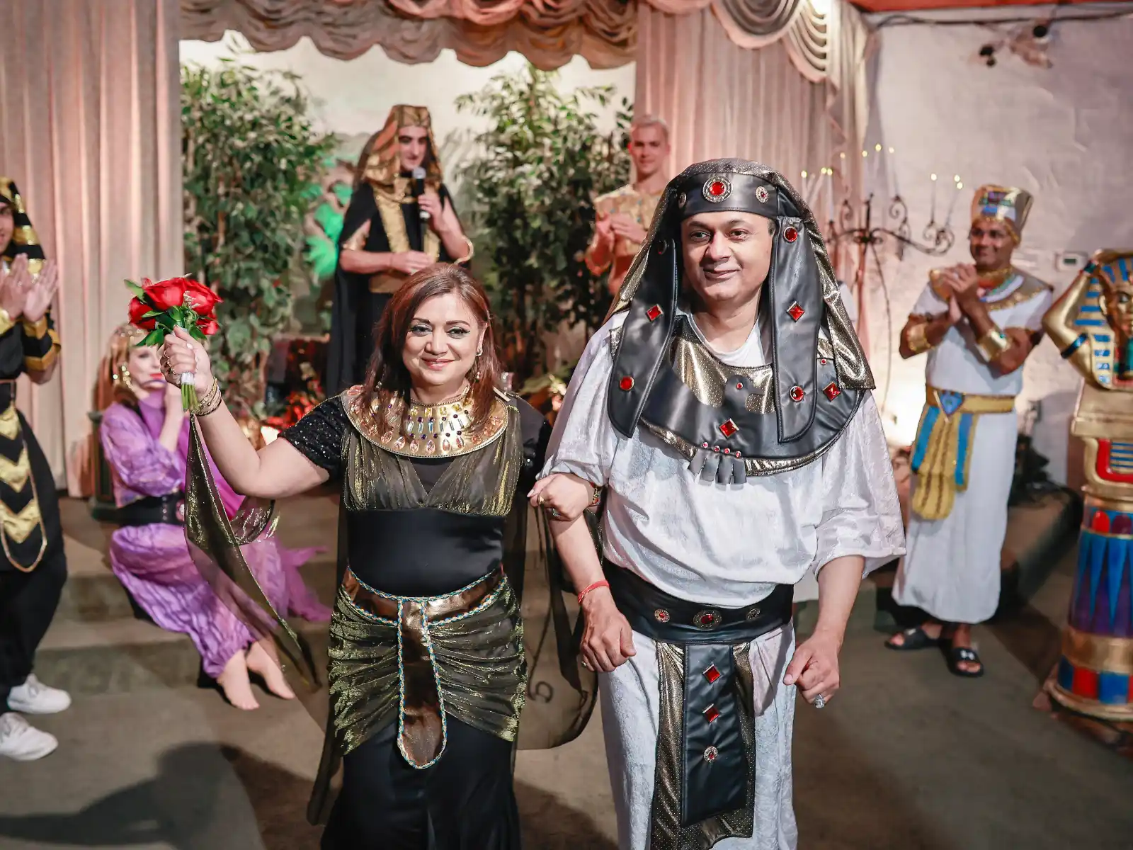 A man and woman in elaborate Egyptian costumes walk forward; the man wears a black and gold Nemes headdress, and the woman holds a bouquet of red roses.