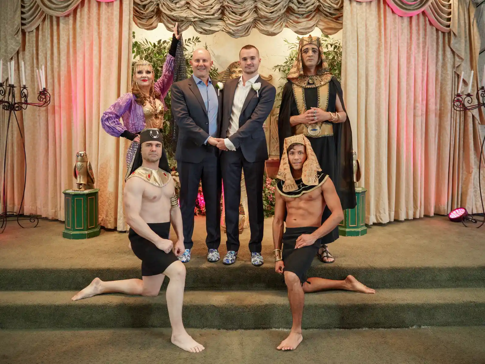 Two men in modern suits stand at the center of a stage, flanked by four performers in Egyptian costumes, including two kneeling men and a woman in purple.