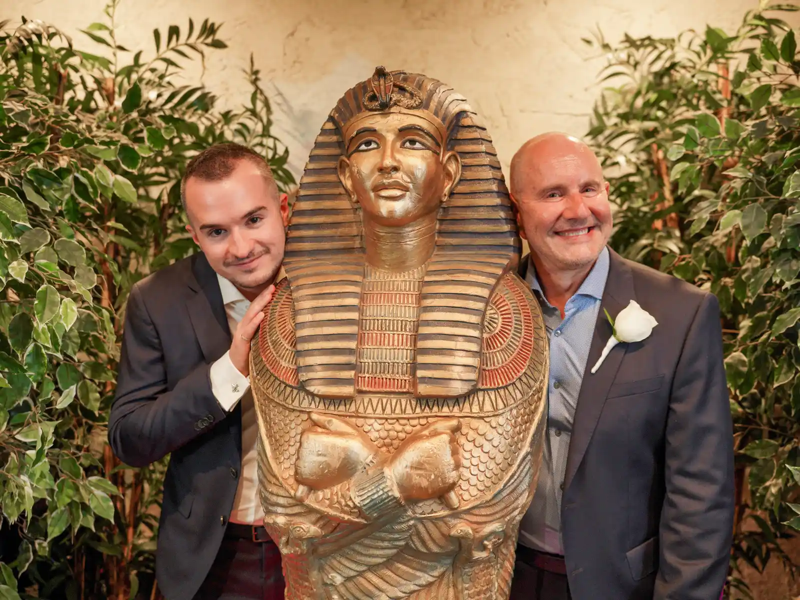 A happy couple in suits posing and leaning against a large, golden King Tutankhamun sarcophagus prop surrounded by greenery.