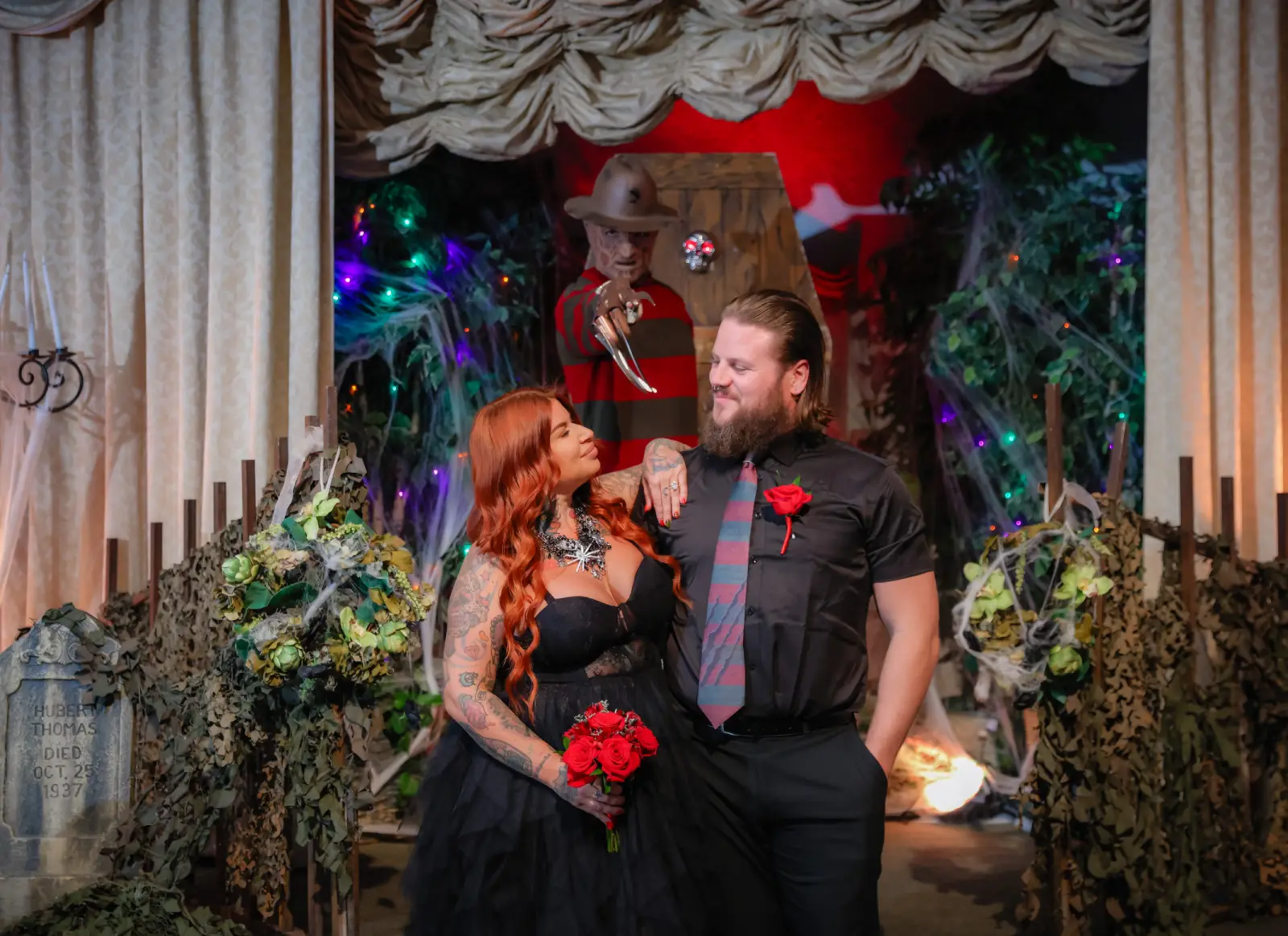 Couple posing in gothic wedding set while Freddy Krueger stands behind in decorated spooky backdrop.