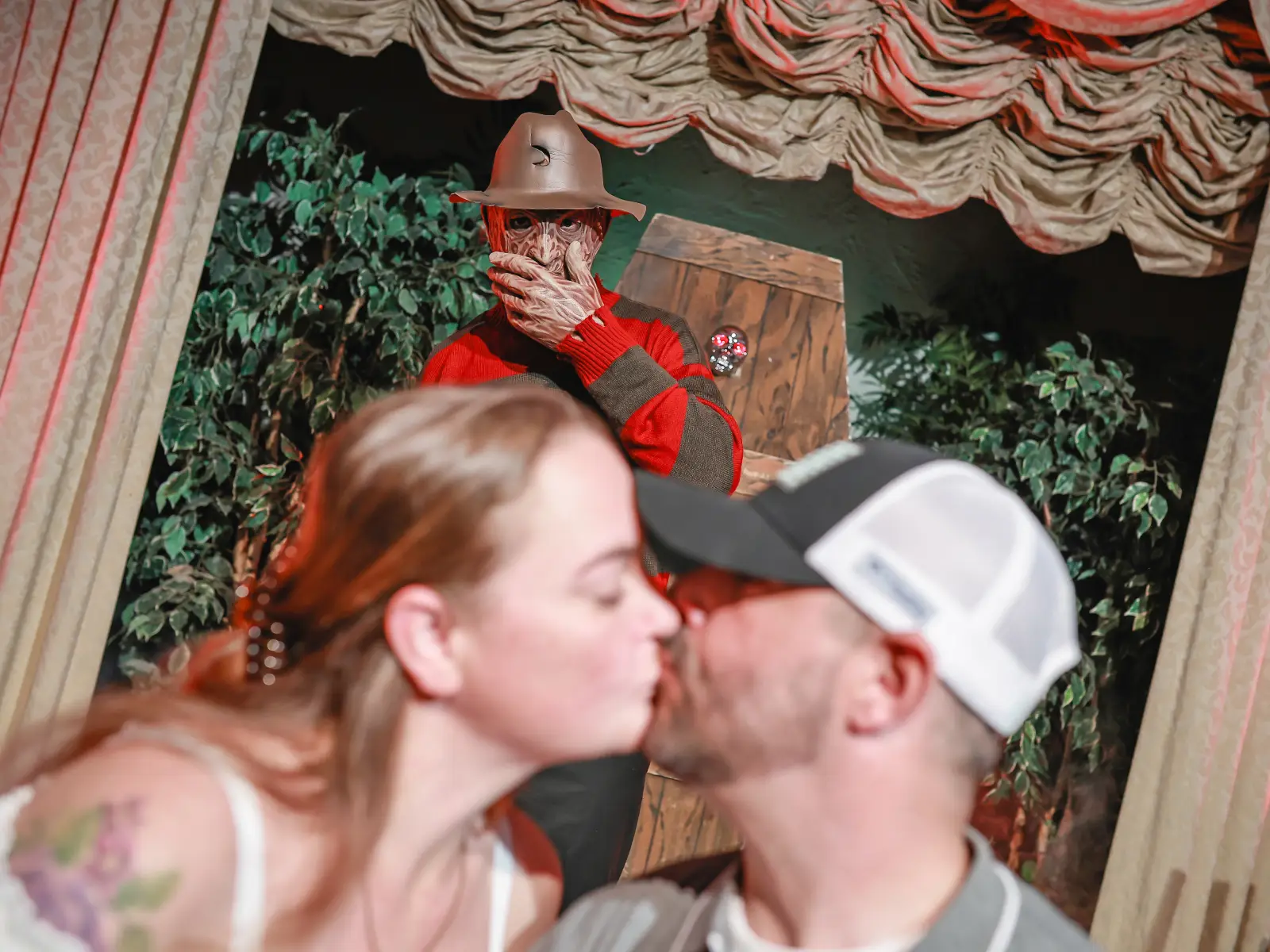 Couple kissing in foreground while Freddy Krueger character watches behind spooky wedding setup.