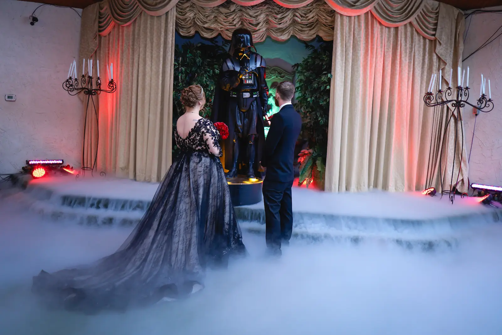 Bride in a black lace gown and groom standing before a Darth Vader officiant in a fog-filled Las Vegas wedding chapel.