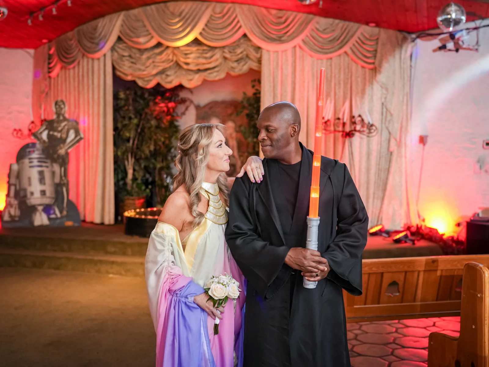 A smiling bride in a red dress and a groom in a yellow martial arts uniform walking down the aisle together after their wedding ceremony, passing a Darth Vader figure standing on the stage.