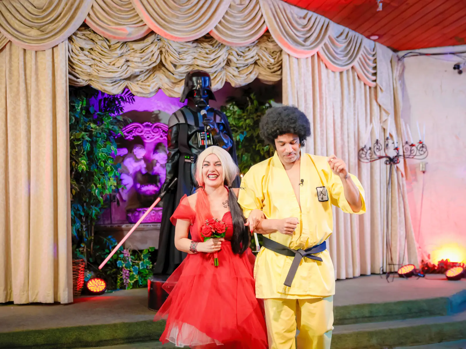 A smiling bride in a red dress and a groom walking down the aisle together after their wedding ceremony, passing a Darth Vader figure standing on the stage.