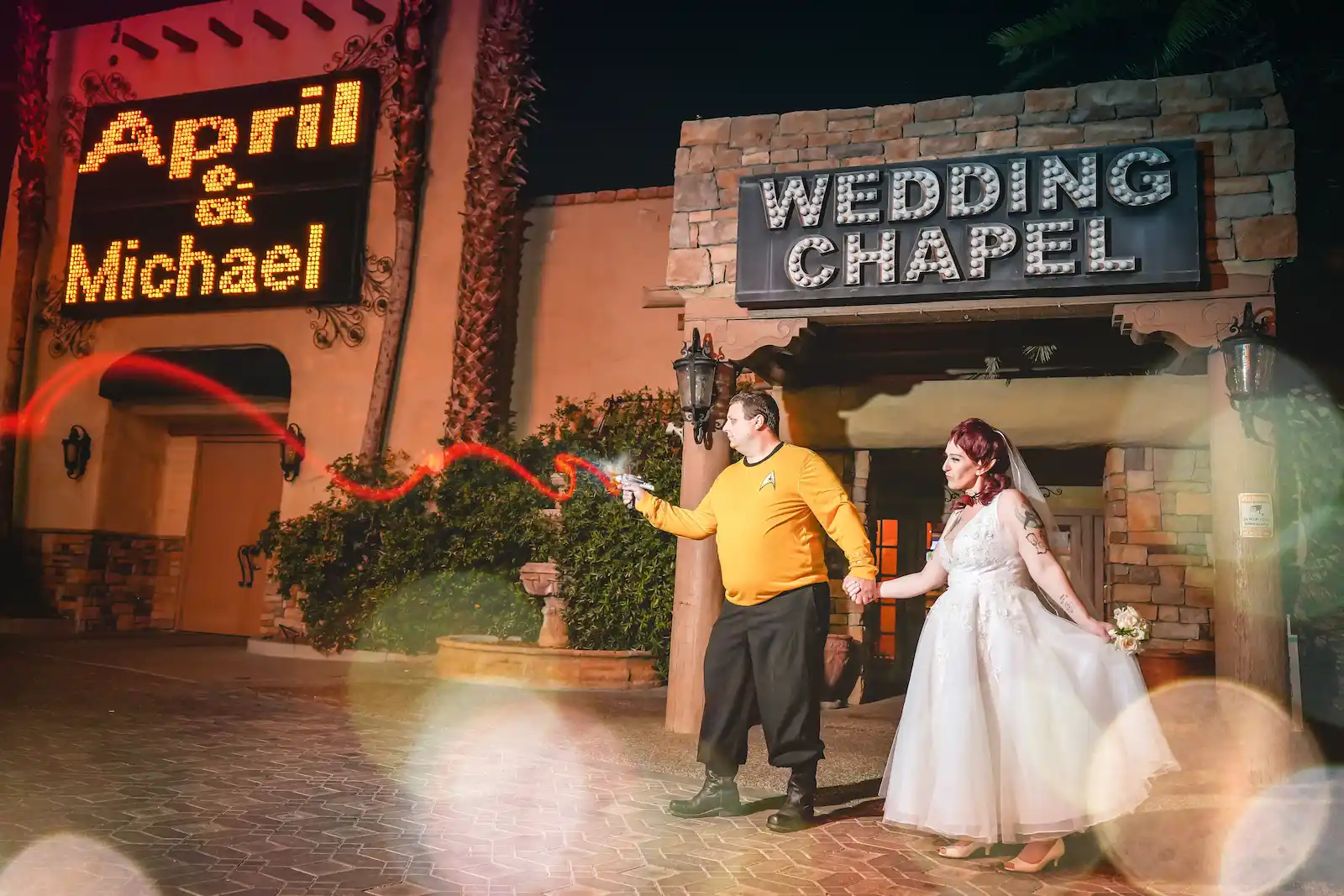 Star Trek-themed groom in a gold uniform firing a toy phaser while holding hands with his bride outside a Las Vegas wedding chapel sign.
