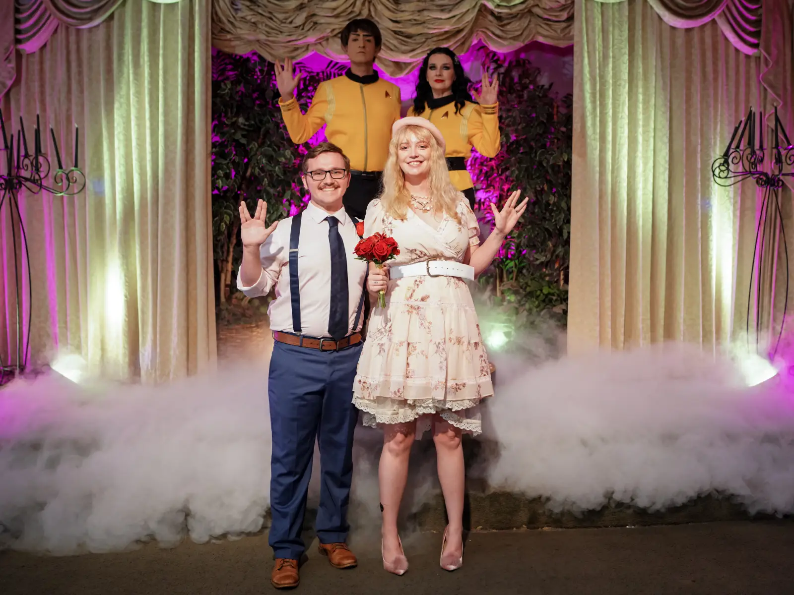 A wedding couple smiling and making the Vulcan salute after their ceremony, with an officiant and another guest in Star Trek uniforms standing on stage behind them.
