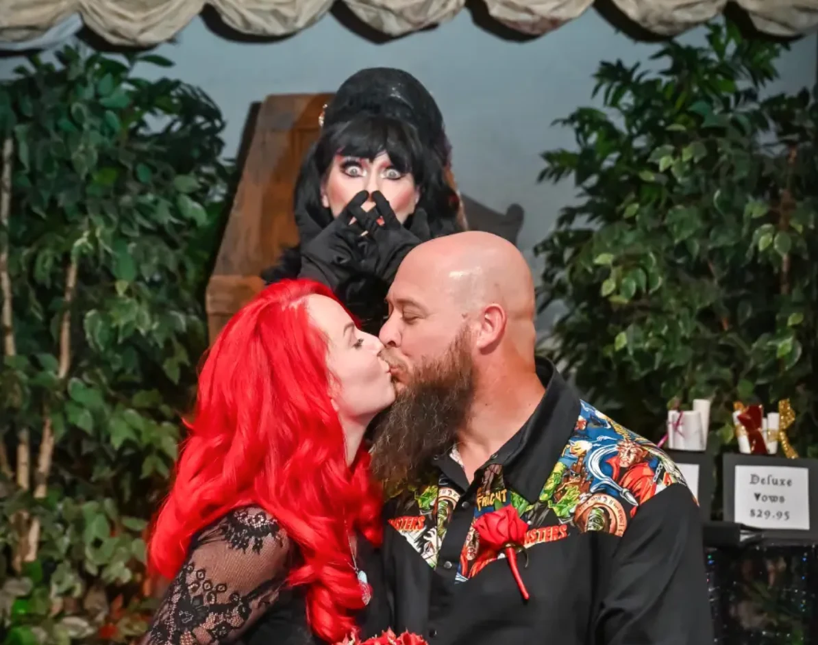 couple kissing during gothic themed wedding ceremony with elvira impersonator behind them