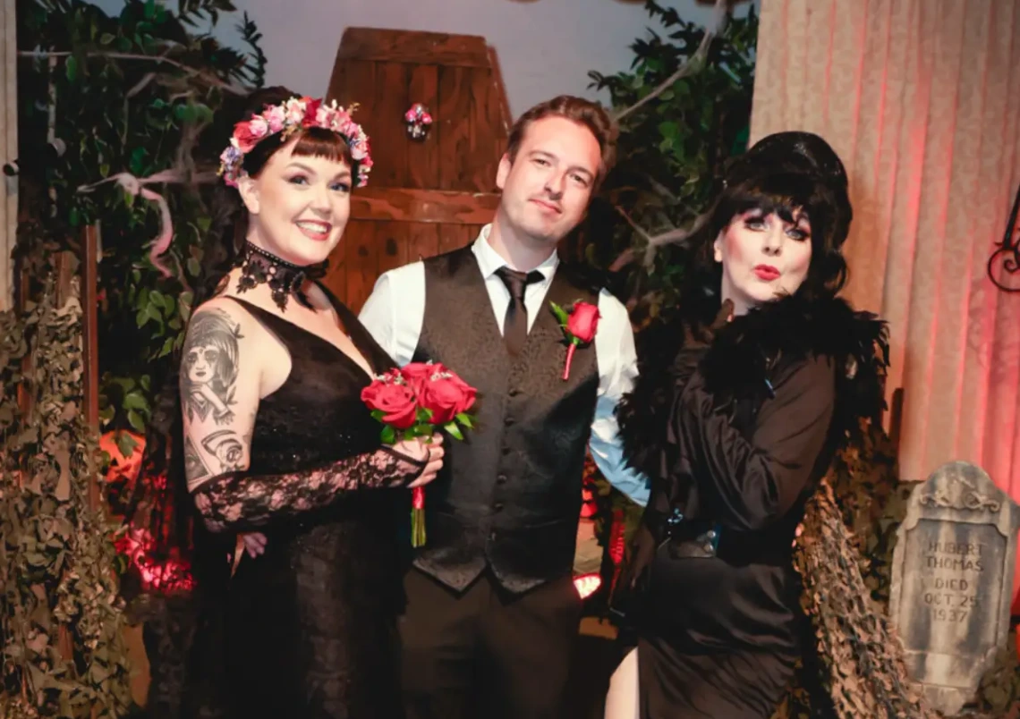 bride groom and elvira impersonator posing together at gothic themed wedding ceremony