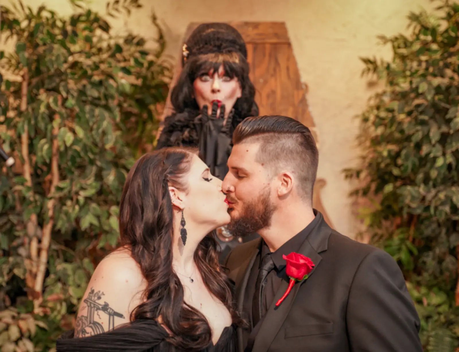 Couple kissing during gothic wedding ceremony while officiant reacts dramatically behind them.